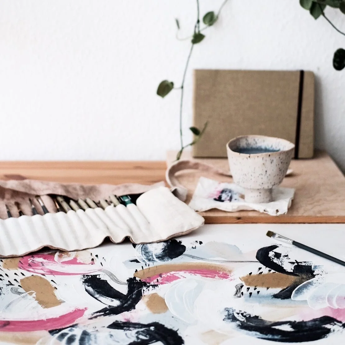 Kunstwerke, Pinsel und eine Tasse auf einem Tisch mit einem Notizbuch im Hintergrund.