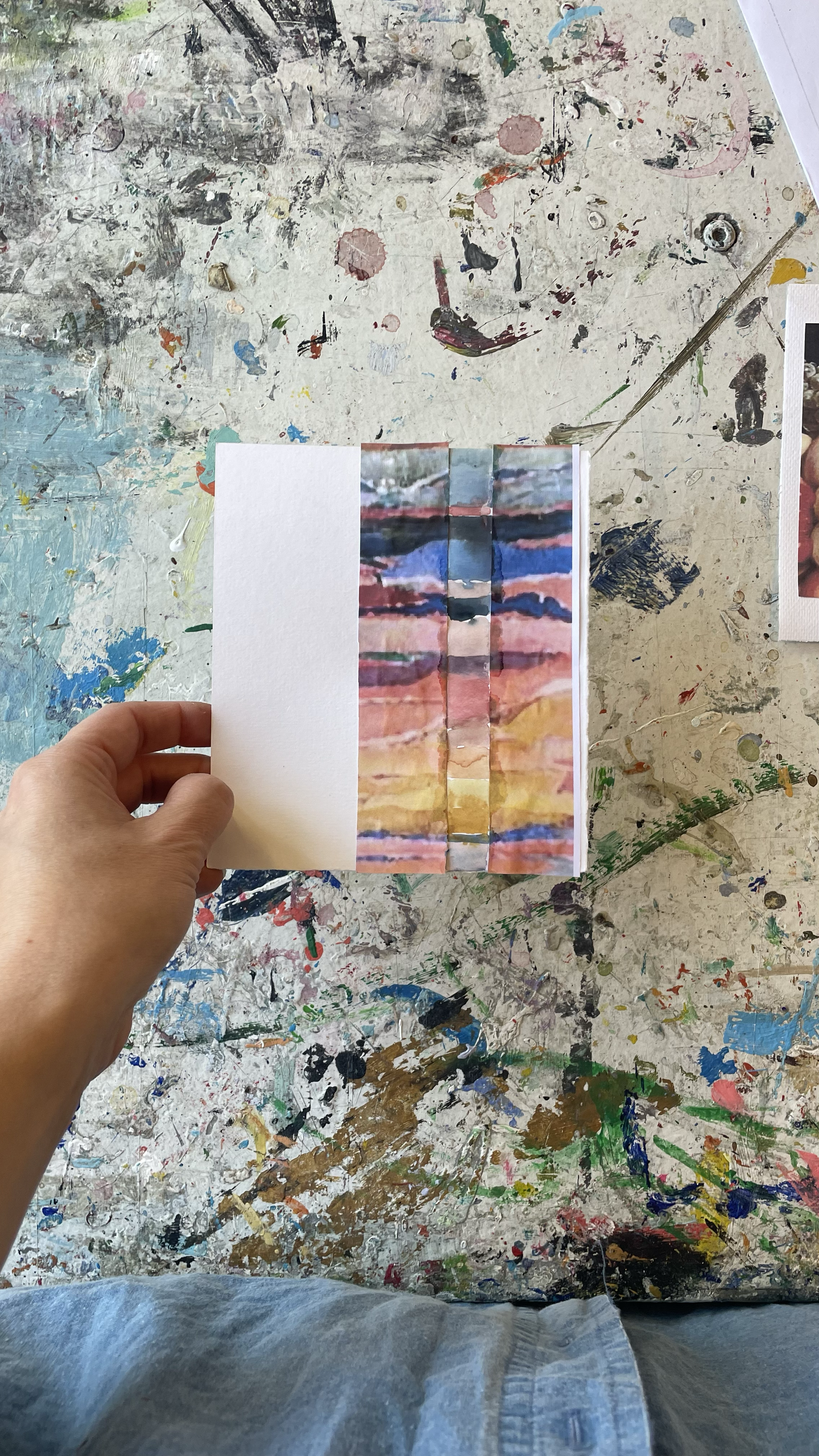 Eine Hand hält eine kleine, bunte, abstrakte Gemäldeseite, die wie ein Sonnenuntergang über Wasser aussieht, vor einem Tisch, der mit Farbflecken und Pinselstrichen bedeckt ist.