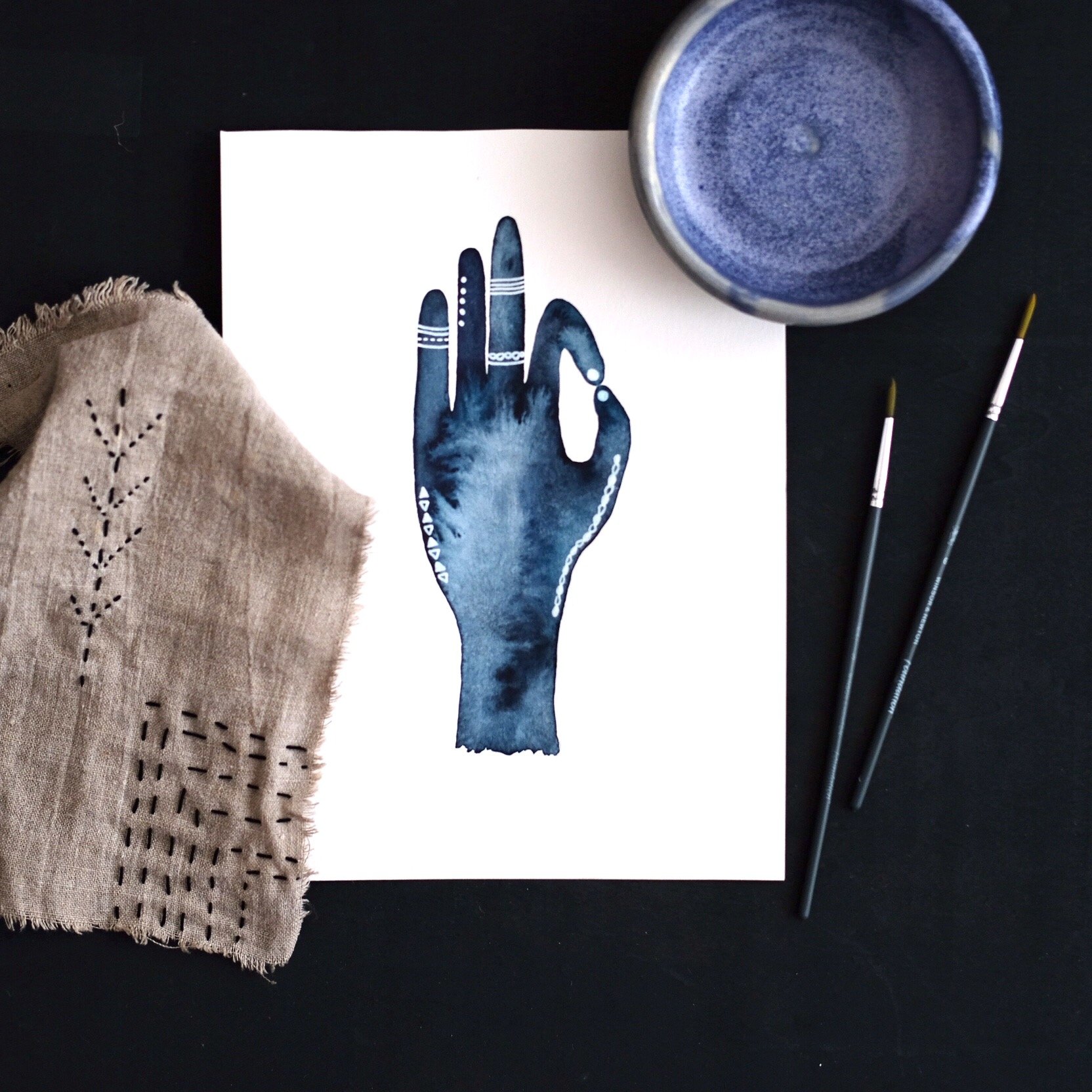 Skizze einer Hand in blauer Aquarelltechnik mit weißen Mustern, daneben ein Tuch mit Stickerei, Pinsel und eine runde Schale auf schwarzem Hintergrund.