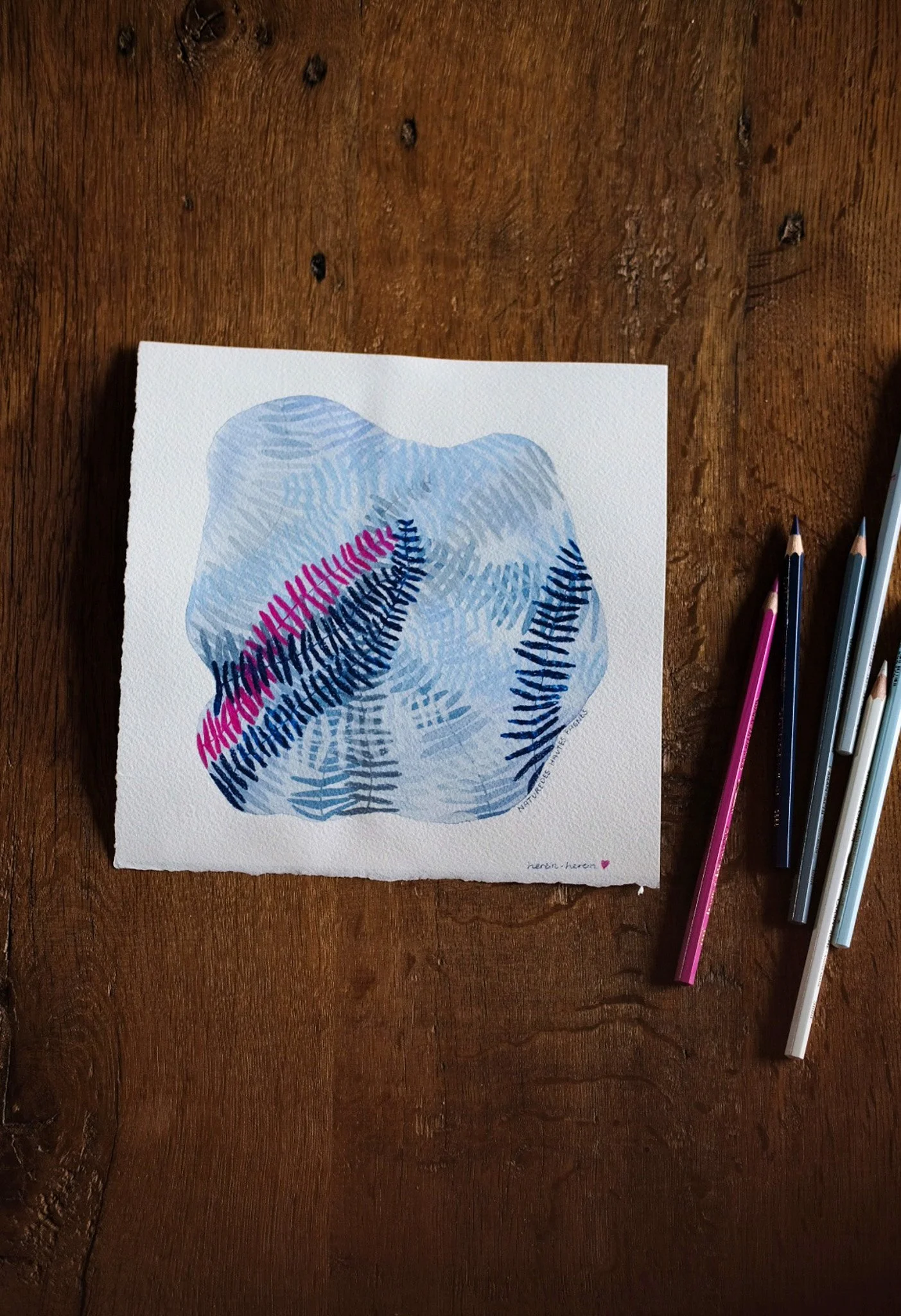 Aquarellbild mit blauen, schwarzen und pinkfarbenen Pinselstrichen auf Papier, umgeben von Buntstiften auf einem Holztisch.
