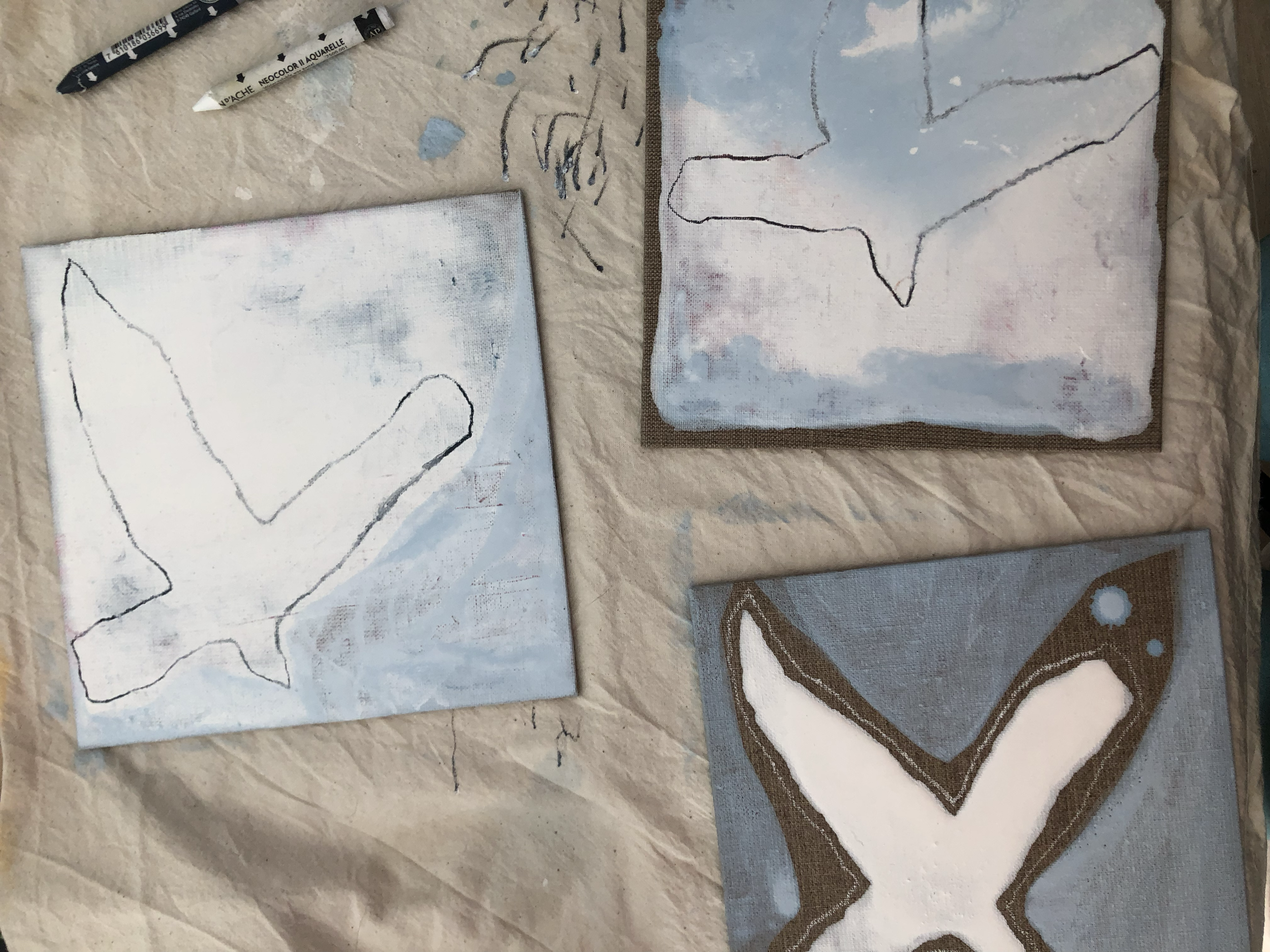 Drei Leinwände, die mit Acrylfarbe bemalt wurden. Zwei Leinwände zeigen eine einfache schwarze Umrisszeichnung auf hellem Hintergrund, eine Leinwand zeigt eine in braun eingerahmte schwarze Umrisszeichnung mit weißem Inneren, auf blauem Hintergrund.