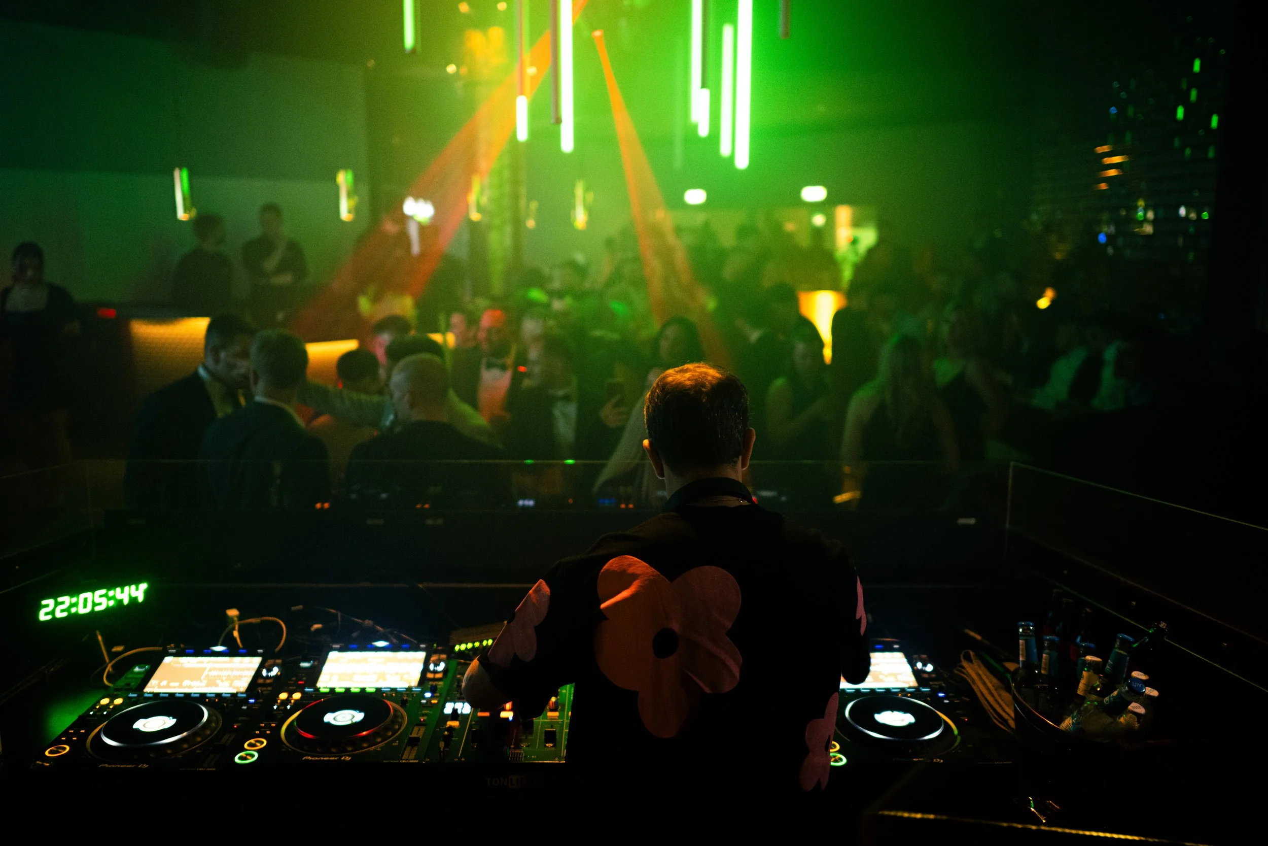 DJ am Mischpult in einer Diskothek mit tanzenden Menschen im Hintergrund, bunte Licht- und Laserstrahlen.