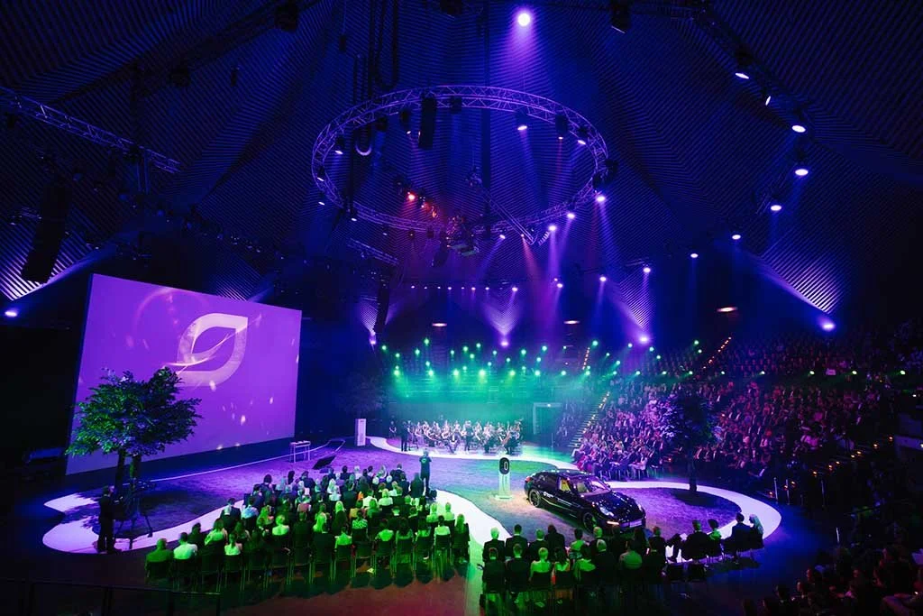 Großes Indoor-Event mit Publikum, einer großen Sound- und Lichtanlage, einer Leinwand mit Logo, einer kleinen Gruppe auf der Bühne, einem schwarzen Auto, grünen Lichteffekten und einer aufgelaufenen Menschenmenge.