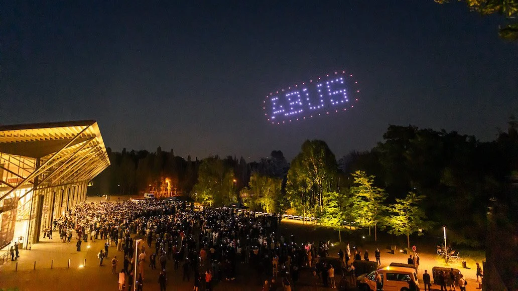 Große Menschenmenge vor einem Gebäude bei Nacht, helle Beleuchtung, Bäume im Hintergrund, Flugzeug aus LED-Lichtern zeigt den Text 'LEUGE' am Himmel.
