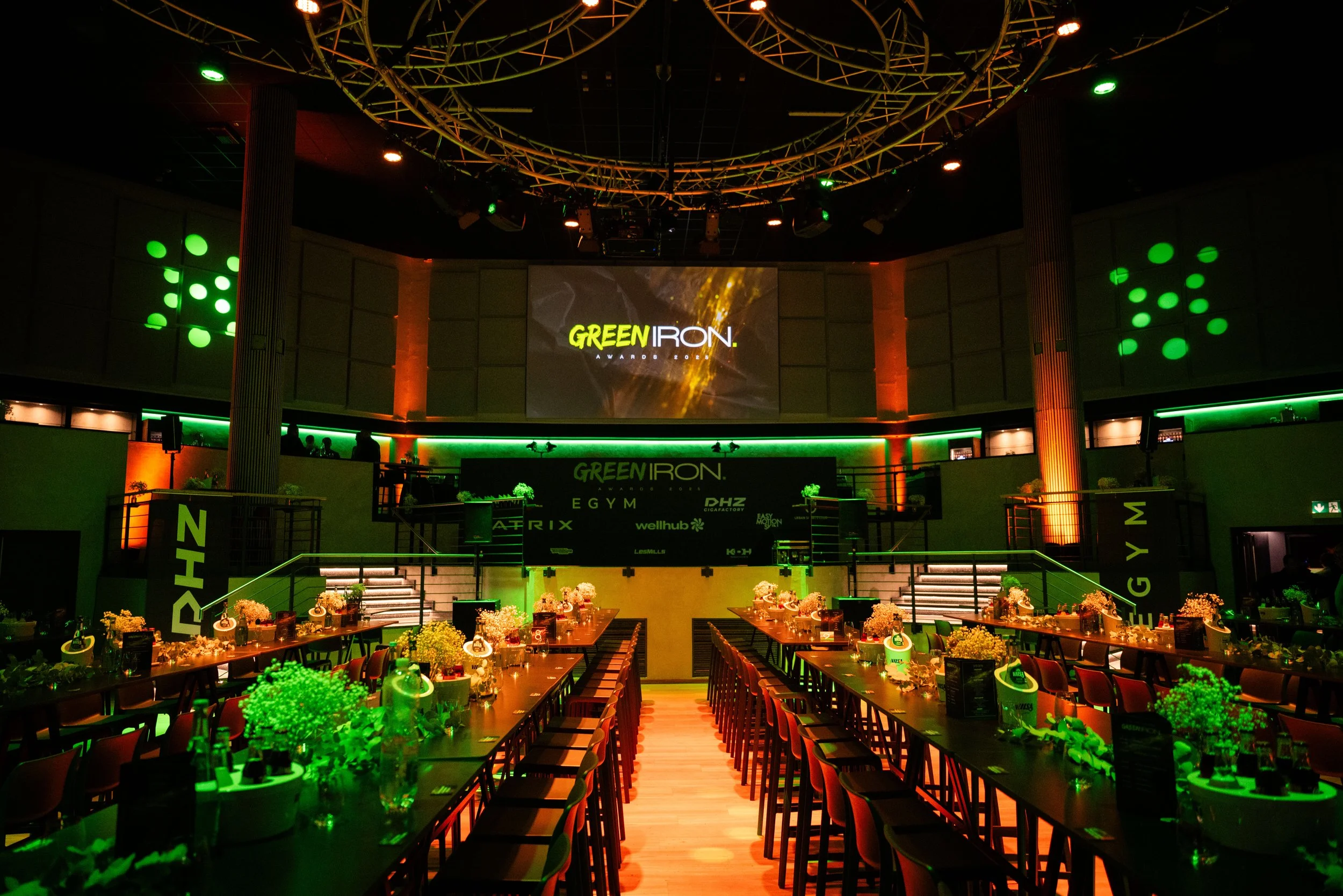 Ein Veranstaltungsraum mit Tischen, dekoriert mit Blumen, auf einer großen Bühne mit einem Bildschirm, der das Logo 'GREEN IRON' zeigt, beleuchtet in grünen und orangefarbenen Lichtern.