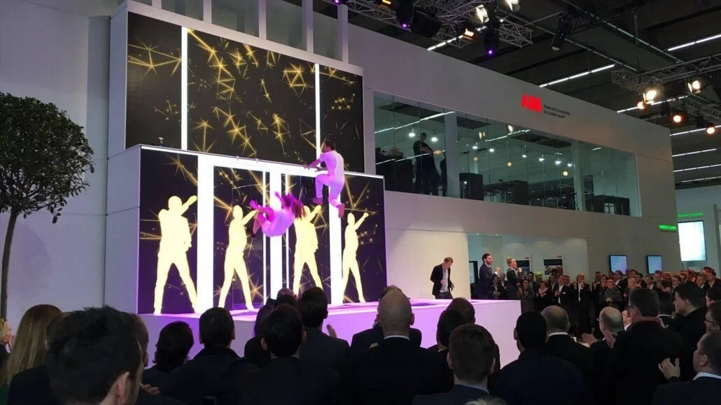 A live performance auf einer Bühne bei einer Messe. Zwei Tänzerinnen in pinken Kostümen tanzen gekonnt, während im Hintergrund leuchtende, gelbe Sternschnuppen auf einem Bildschirm zu sehen sind. Das Publikum schaut zu.