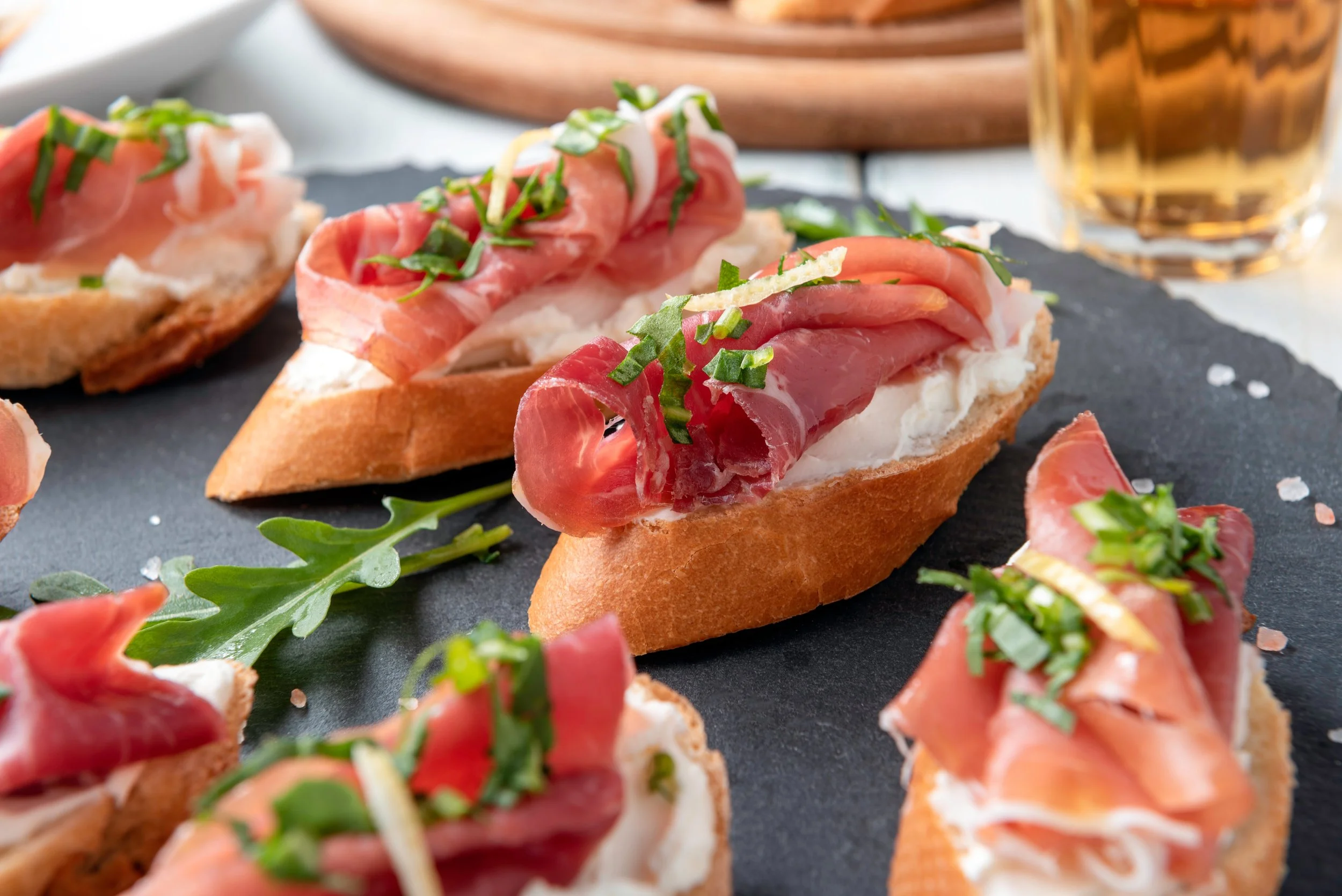 Bunte geschnittene Bruschetta mit Schinken, Sprossen und Kräutern auf einer schwarzen Platte, im Hintergrund ein Glas Bier.