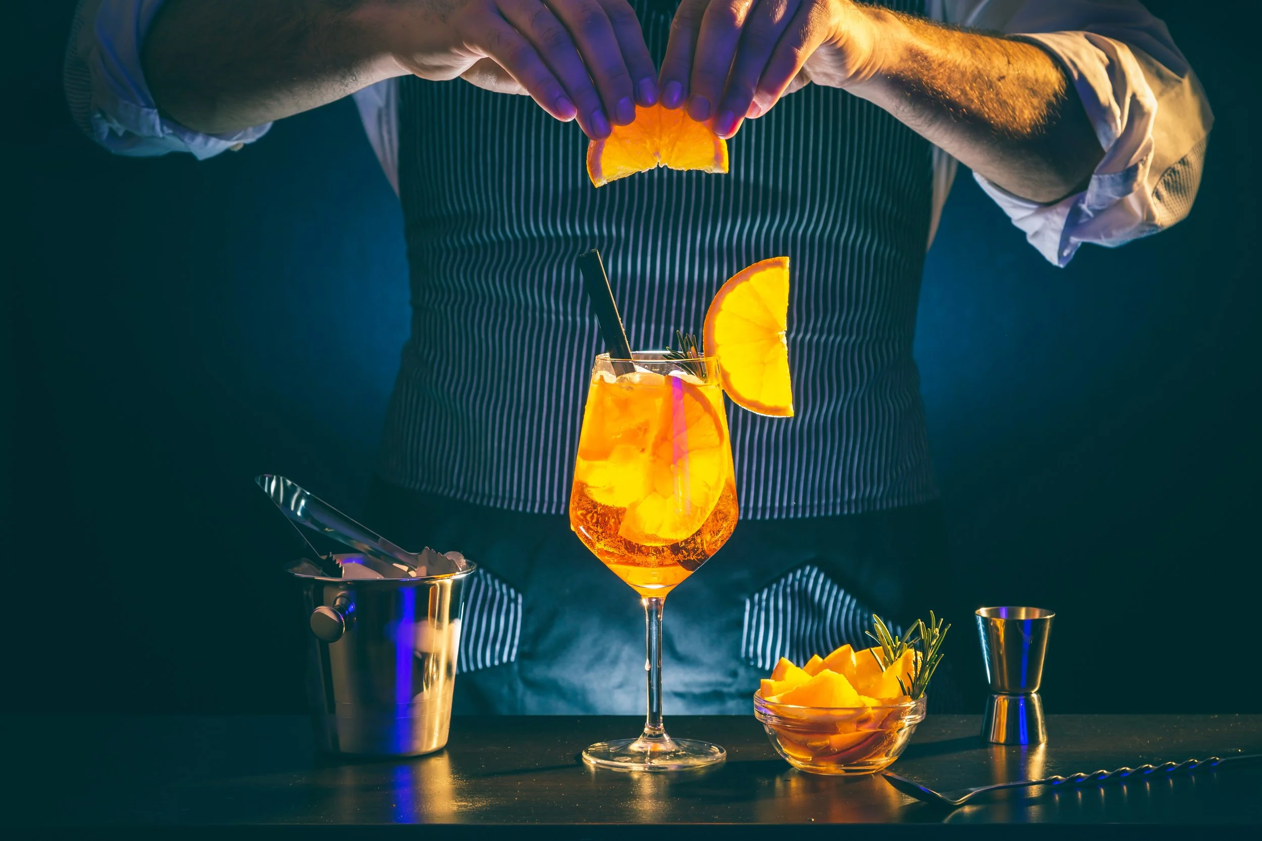 Barmann bereitet einen orangefarbenen Cocktail mit Orangenscheiben und Rosmarin in einem Weinglas vor.
