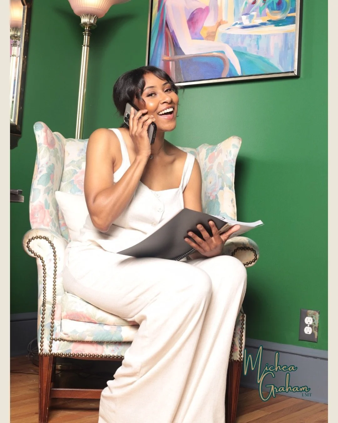 A woman in white pajamas sitting on a floral upholstered armchair, smiling and talking on a cellphone, holding a notebook, in a room with green walls, a painting, and a floor lamp.