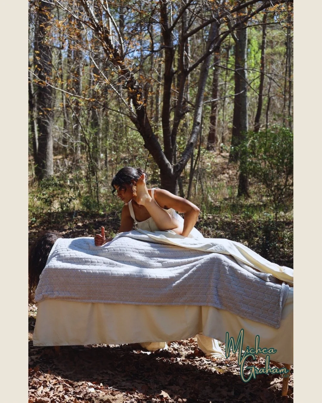 A woman giving a massage on a massage table outdoors in a forest. She is wearing a sleeveless top and is giving a thumbs-up while looking at another person lying on their stomach under a blanket. There are trees and sunlight in the background. The ph