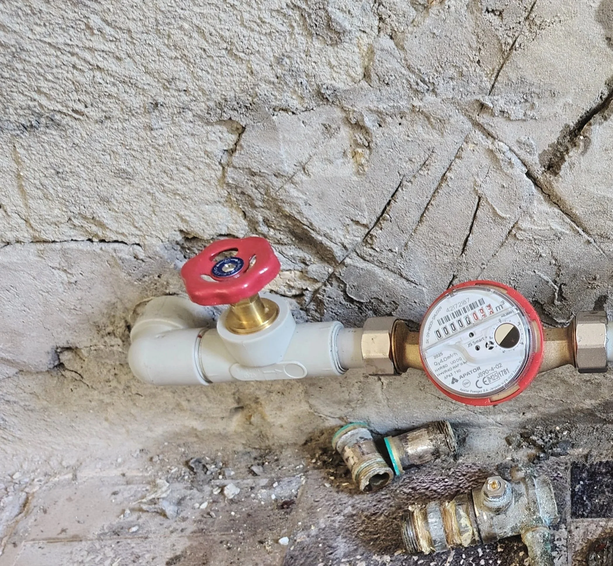 Water meter connected to a white pipe with a red-handled valve, against a stone wall, with additional plumbing components on the ground.