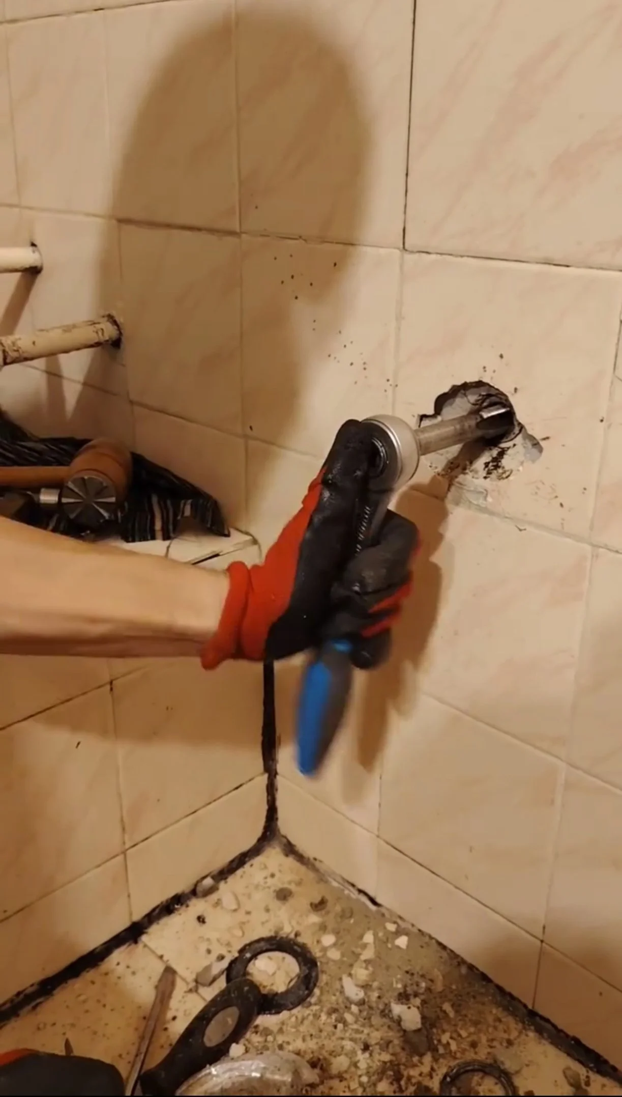 A person in black gloves using a power drill to remove a pipe from a tiled wall in a bathroom.
