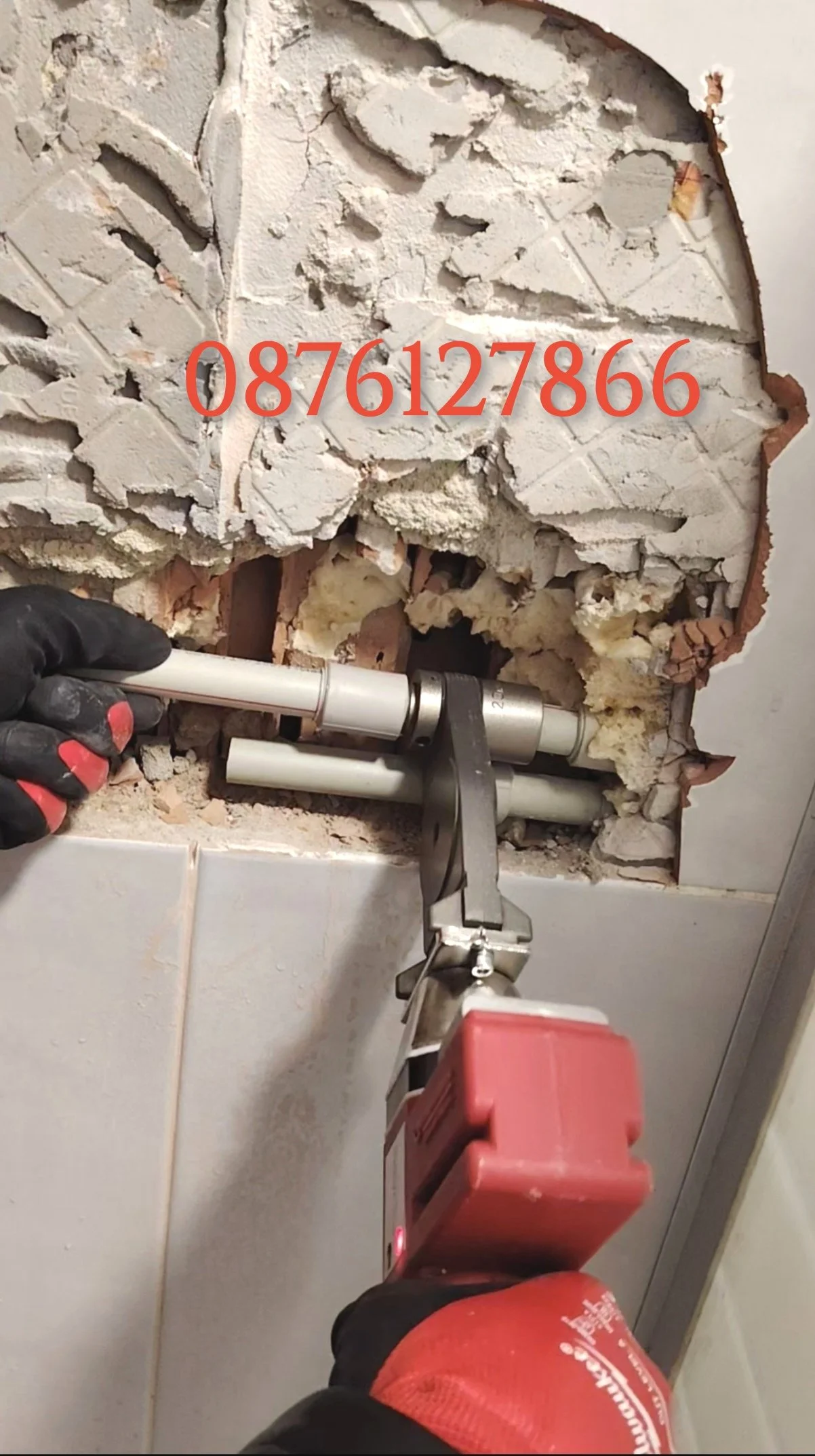 A person wearing black gloves using a power drill to remove or repair plumbing pipes through a wall with exposed drywall and insulation. The wall has some large holes and cracks, with debris around the area.