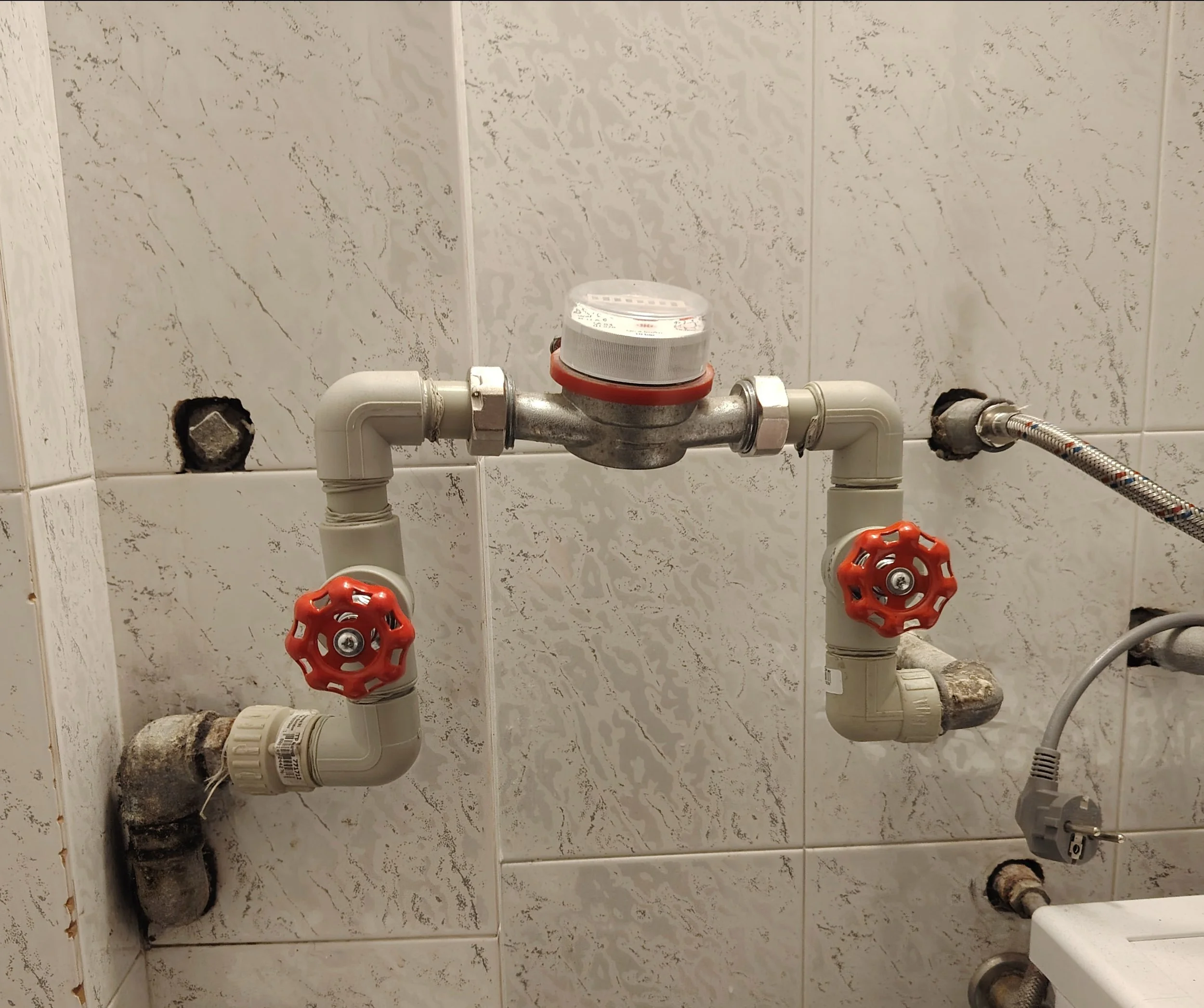 Water meter connected to plumbing with two red valves on pipes in a tiled wall.