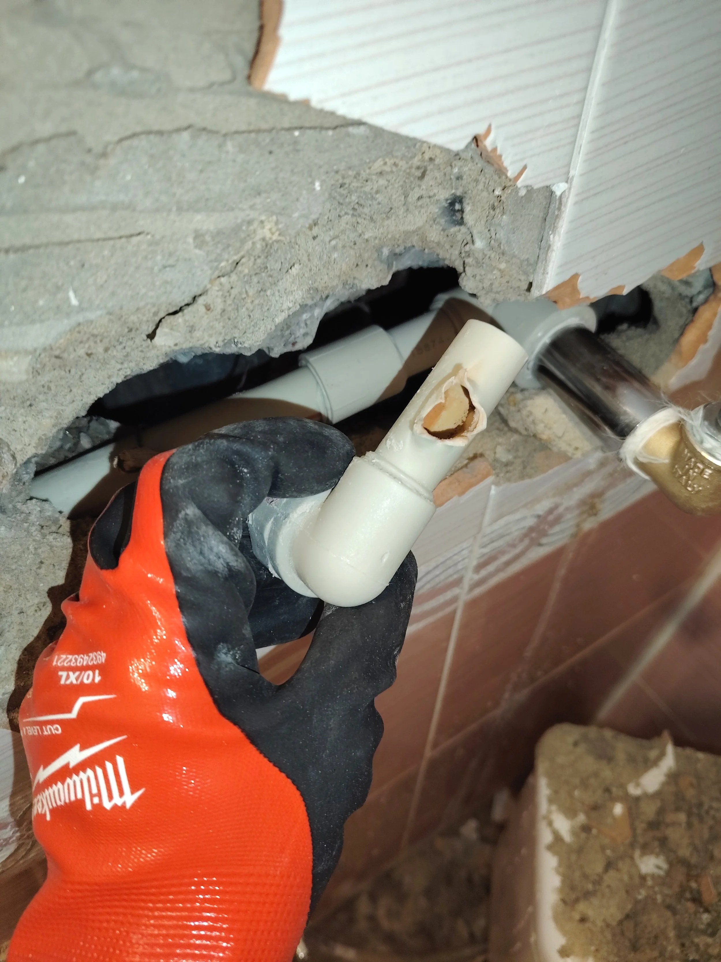 A person wearing a black and orange work glove holding a damaged white PVC pipe with a hole in it, near a hole in the wall with visible inner piping and construction debris.