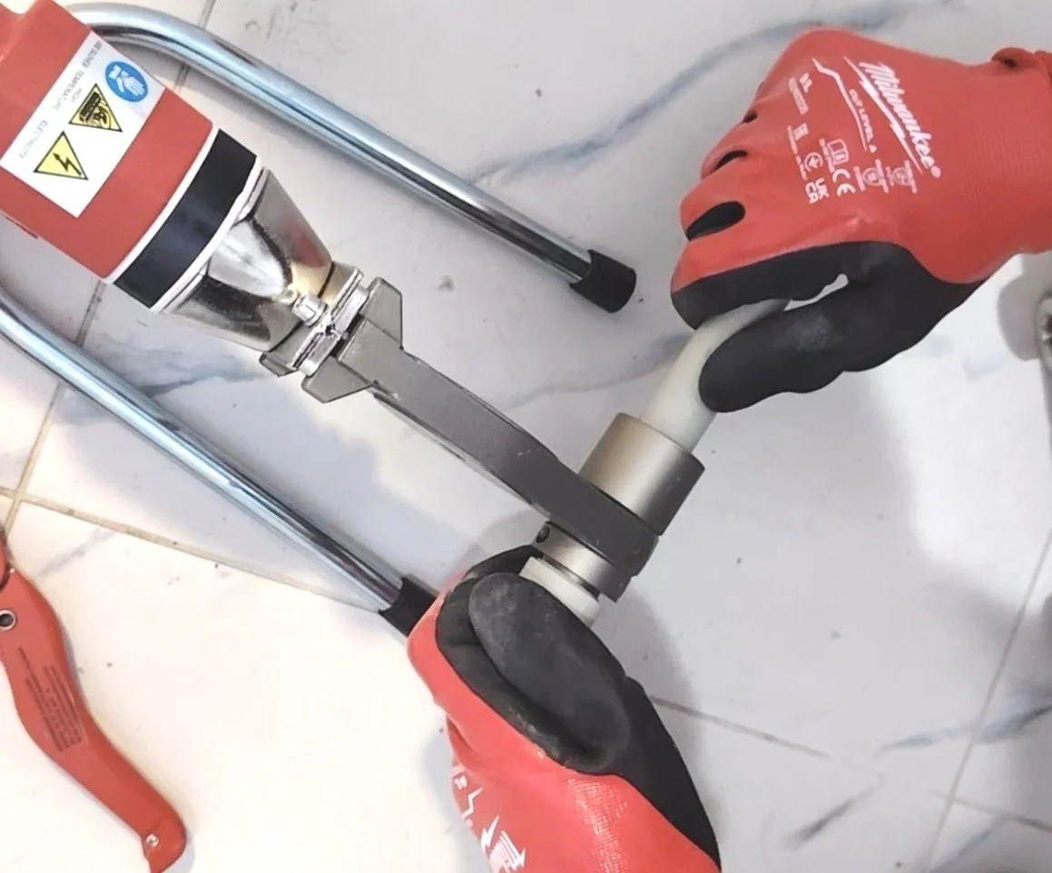 A worker wearing red gloves is installing or repairing a metallic pipe fitting, using a wrench, on a white tiled floor.