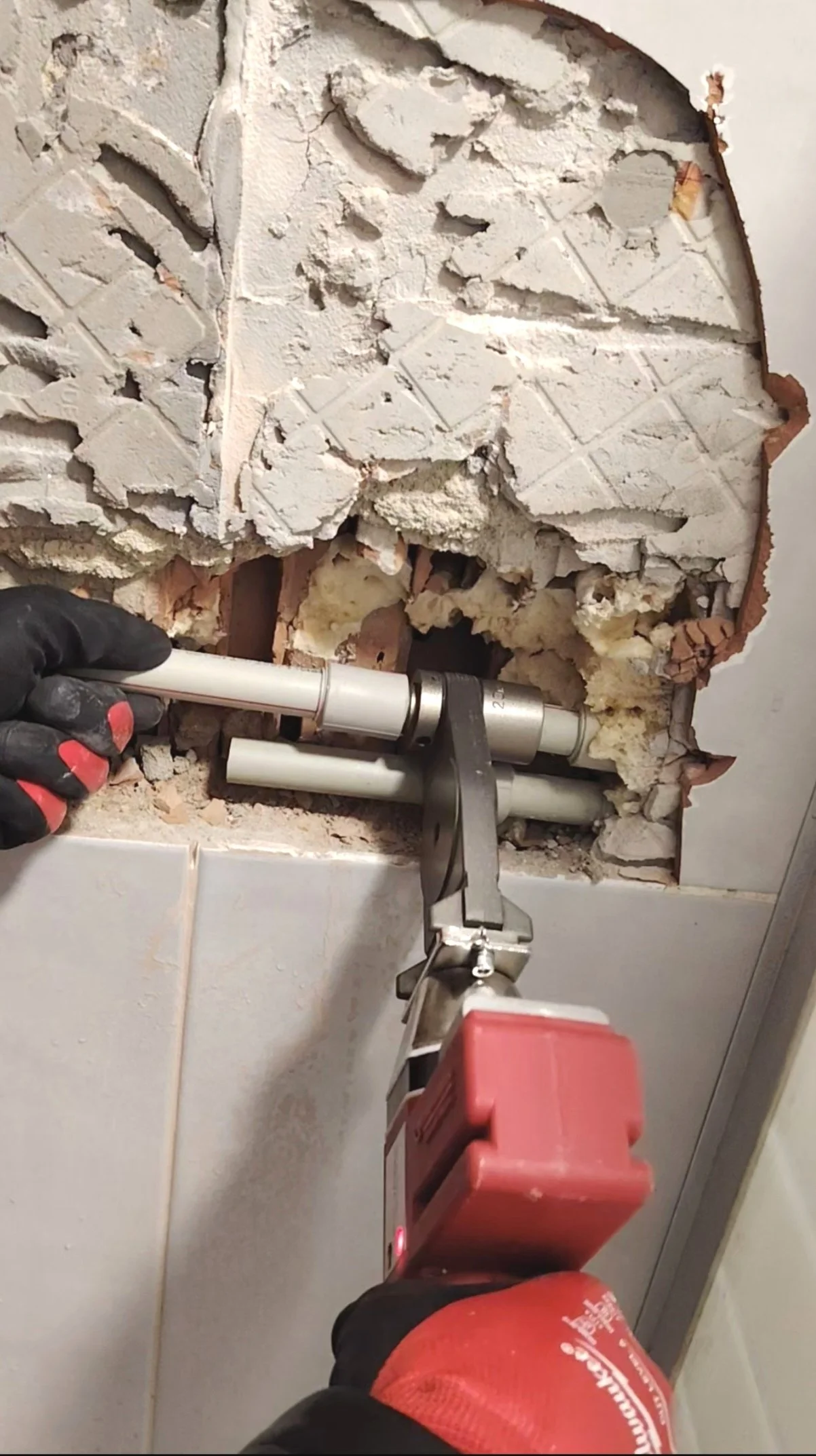 Person using a power tool to remove or repair plumbing behind a wall with exposed insulation and broken tiles.