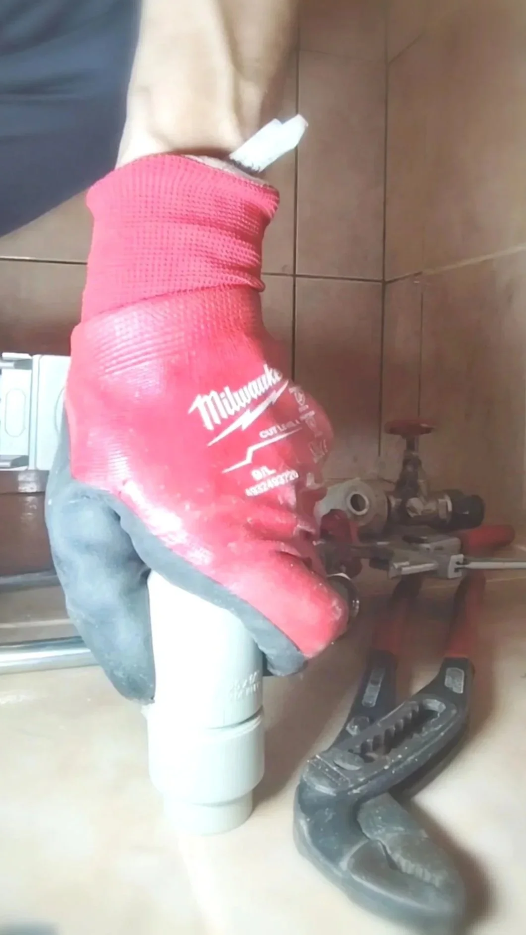 A close-up of a construction worker's gloved hand, wearing a red Milwaukee glove, holding a pneumatic tool in a workshop or construction site with various tools on a table and a brown tiled wall in the background.