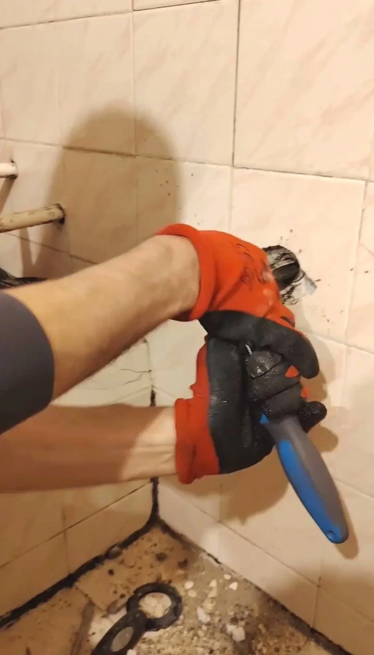 A person wearing orange gloves and holding a blue utility knife is cutting a black pipe inside a tiled bathroom corner.