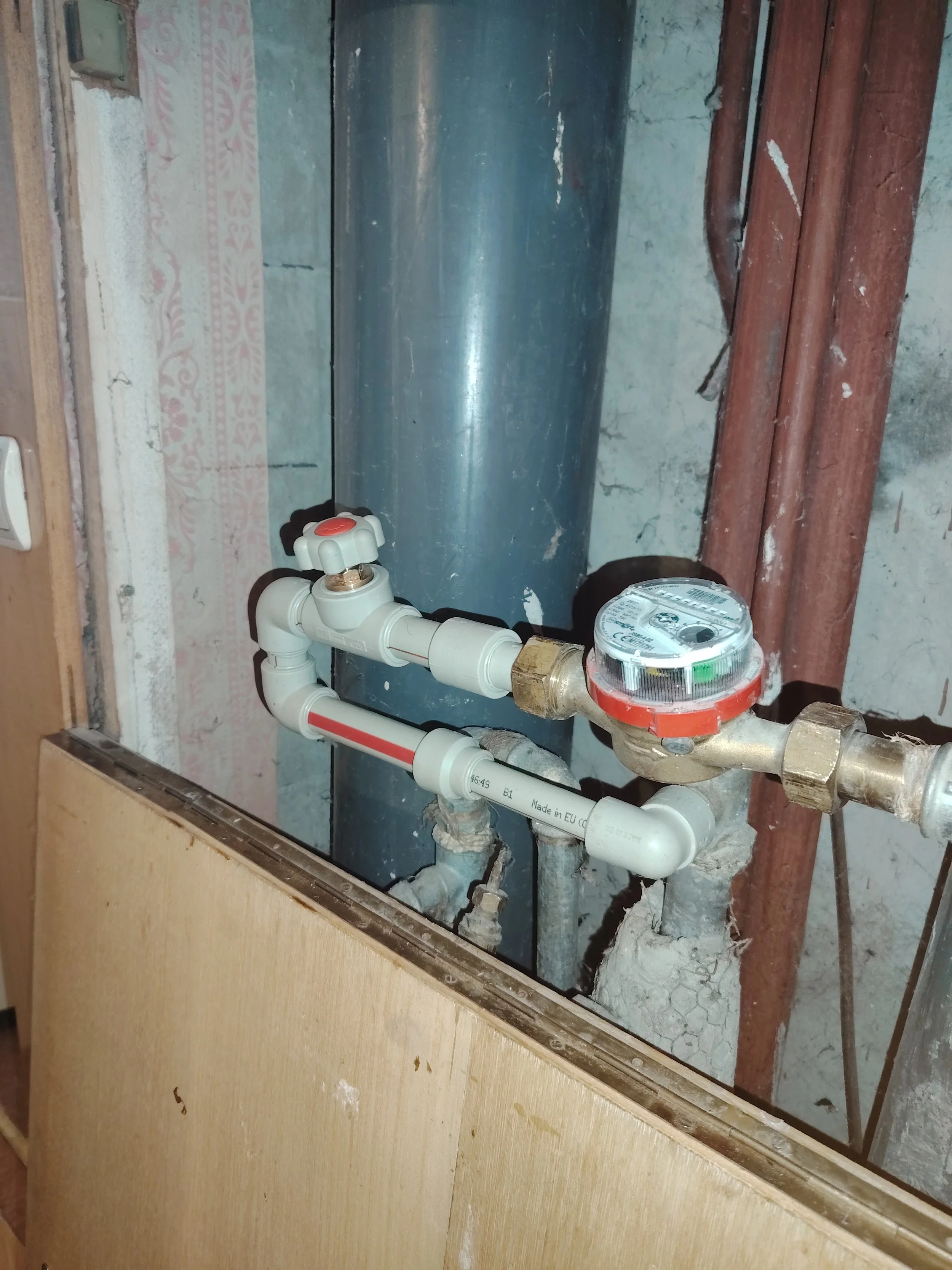 A water meter and piping system behind a cut-out in a wall, with some rusted pipes and a water softener or filter. The background shows a rough wall surface with some peeling paint.