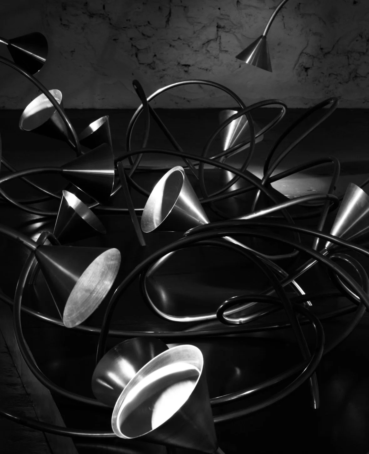 An abstract sculpture made of intertwined metallic cones and rods, displayed in a gallery with a textured wall background.