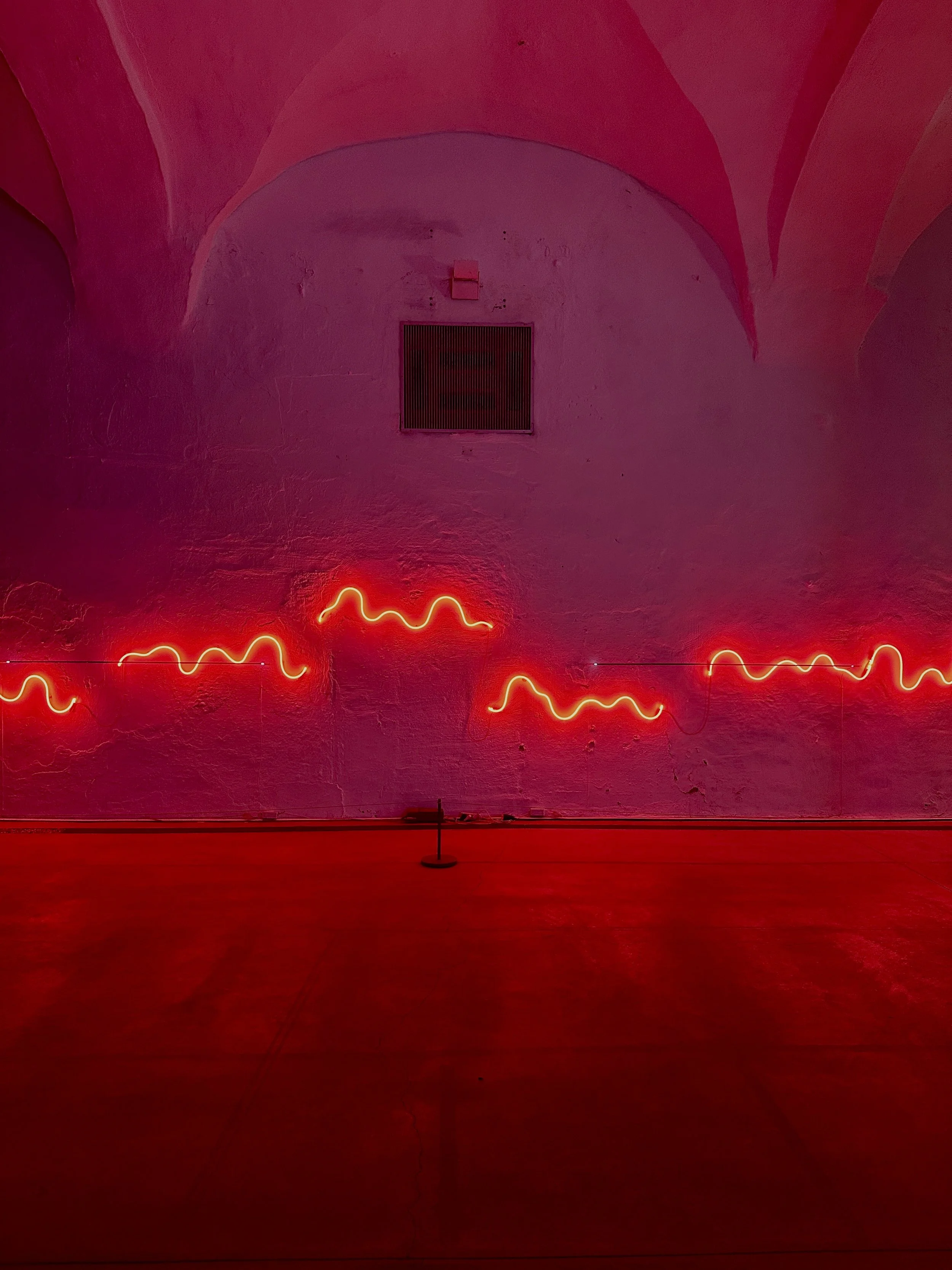 Red neon wavy light installation on a textured wall inside a building with arched ceiling.