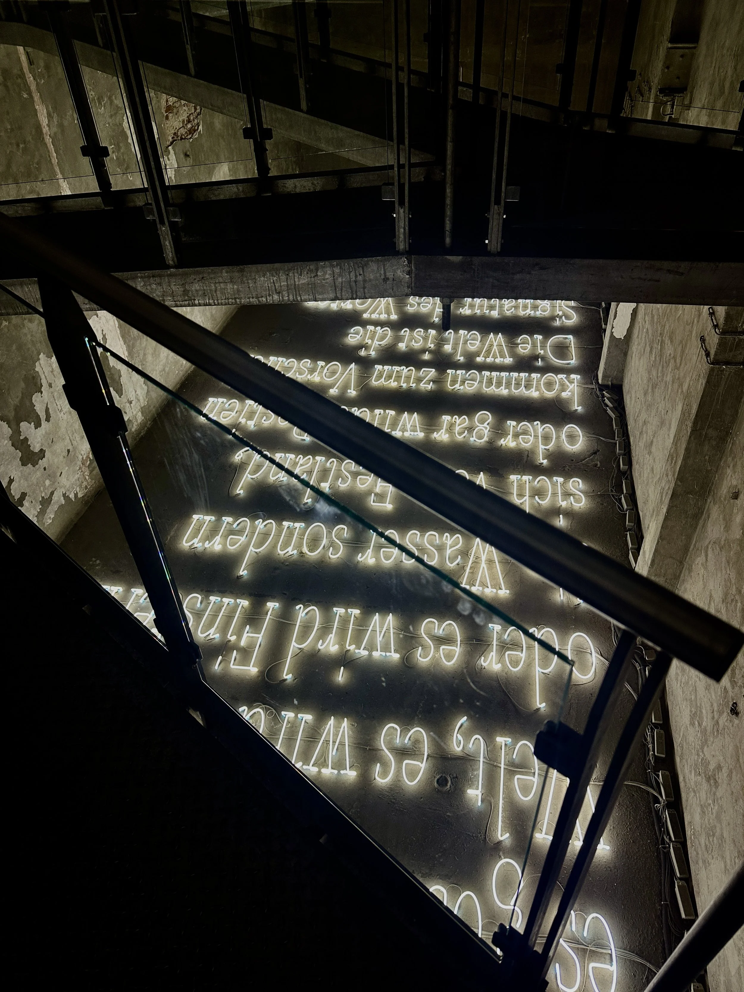 Neon sign with white text on a dark surface, visible from the ceiling, featuring a quote about wine and wisdom with surrounding industrial-style walls and railings.