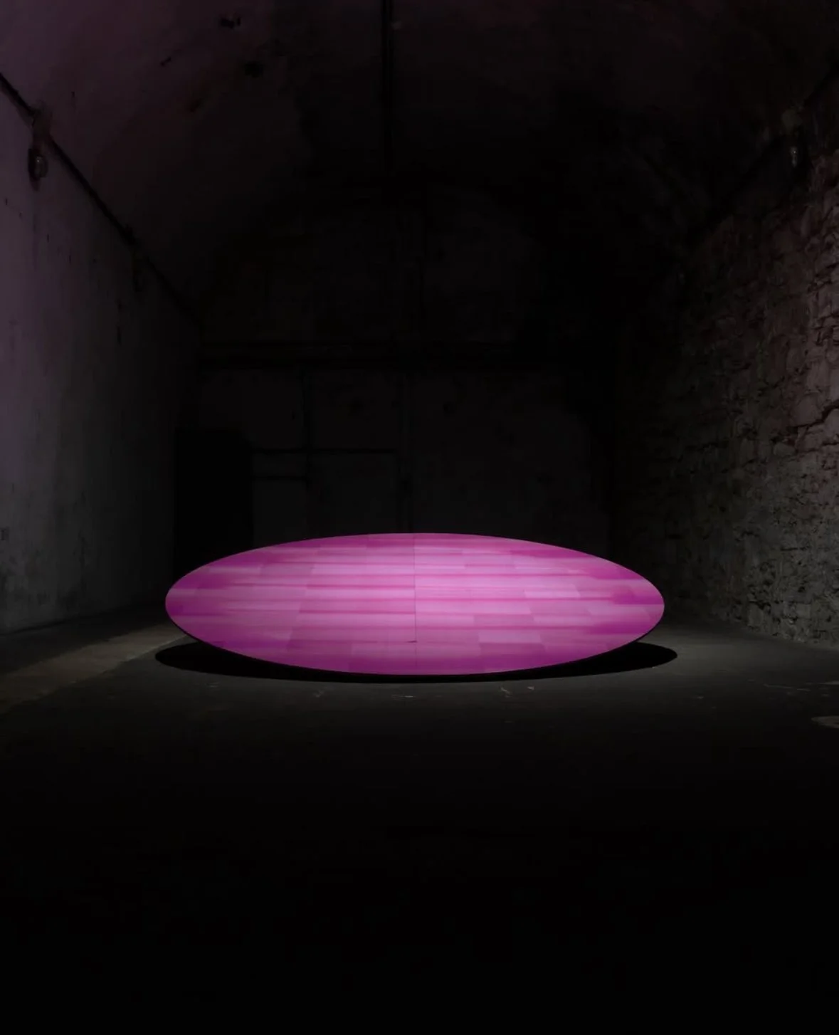 An oval-shaped, glowing pink art installation in a dark, industrial-style gallery with brick and concrete walls and a concrete floor, casting a shadow on the floor.