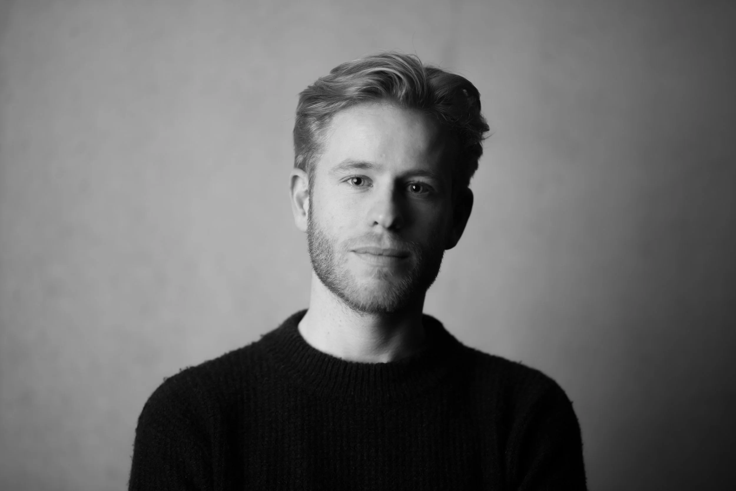 Zwart-wit portret van een man met lichte huid, lichte ogen, lichtbruin haar en een volle baard, gekleed in een zwarte trui, met een neutrale uitdrukking, kijkend naar de camera.