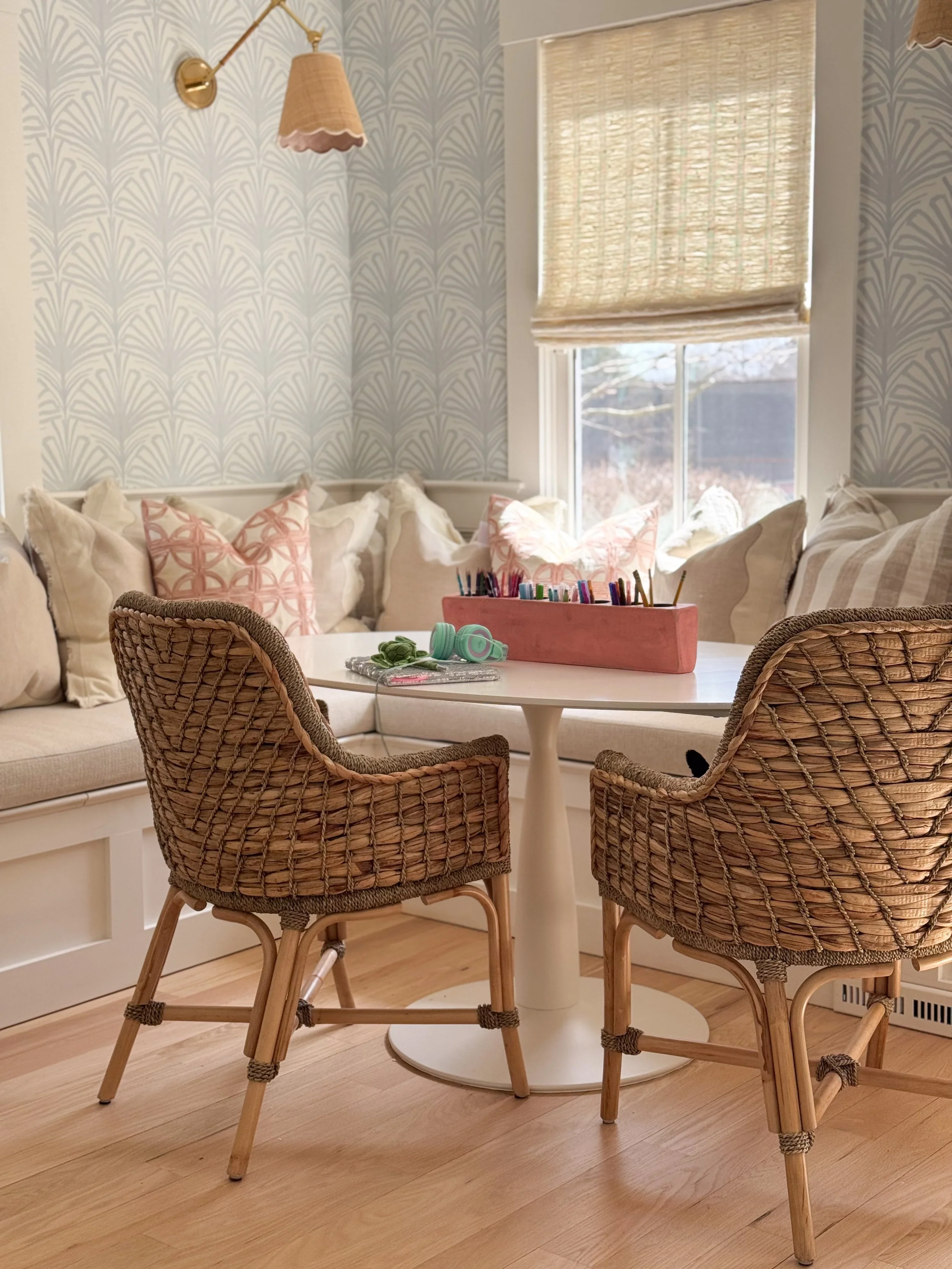 Hardwood floor refinishing in a Scituate dining nook with a white round table, wicker chairs, a built-in bench with cream and pink pillows, patterned wallpaper, and a brass wall sconce