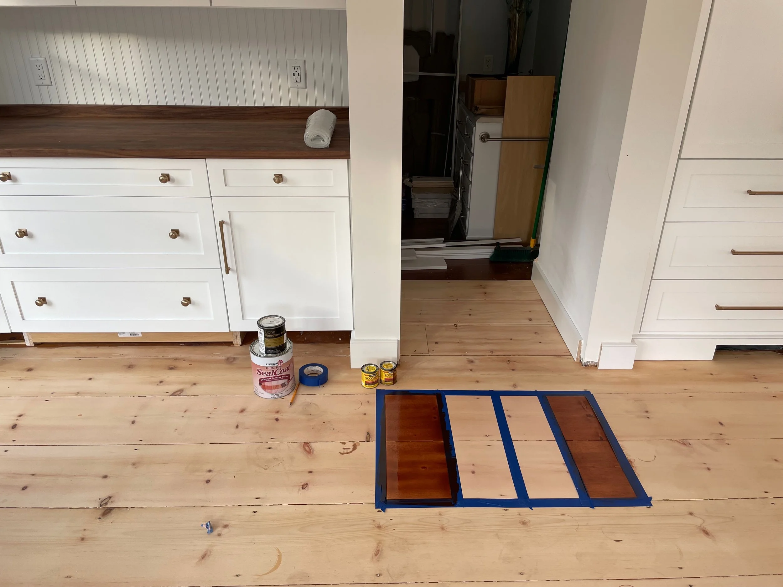 Hardwood floor refinishing in progress in a Duxbury kitchen, with stain samples, painter’s tape, and painting supplies laid out on the wood flooring.