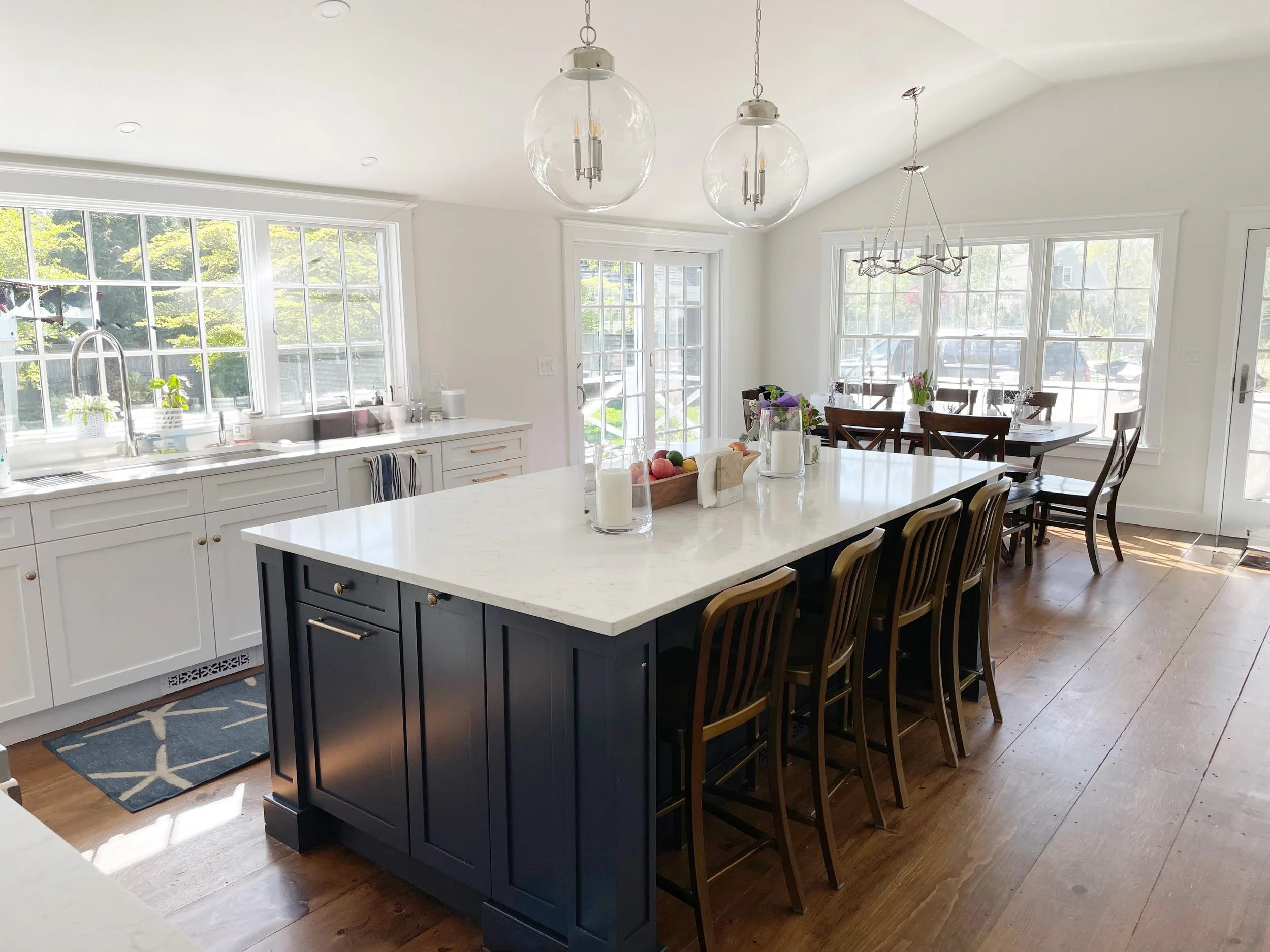 Professional wide pine refinishing in a Duxbury kitchen with white cabinets, a black island, expansive windows, and a warm wood dining setup.