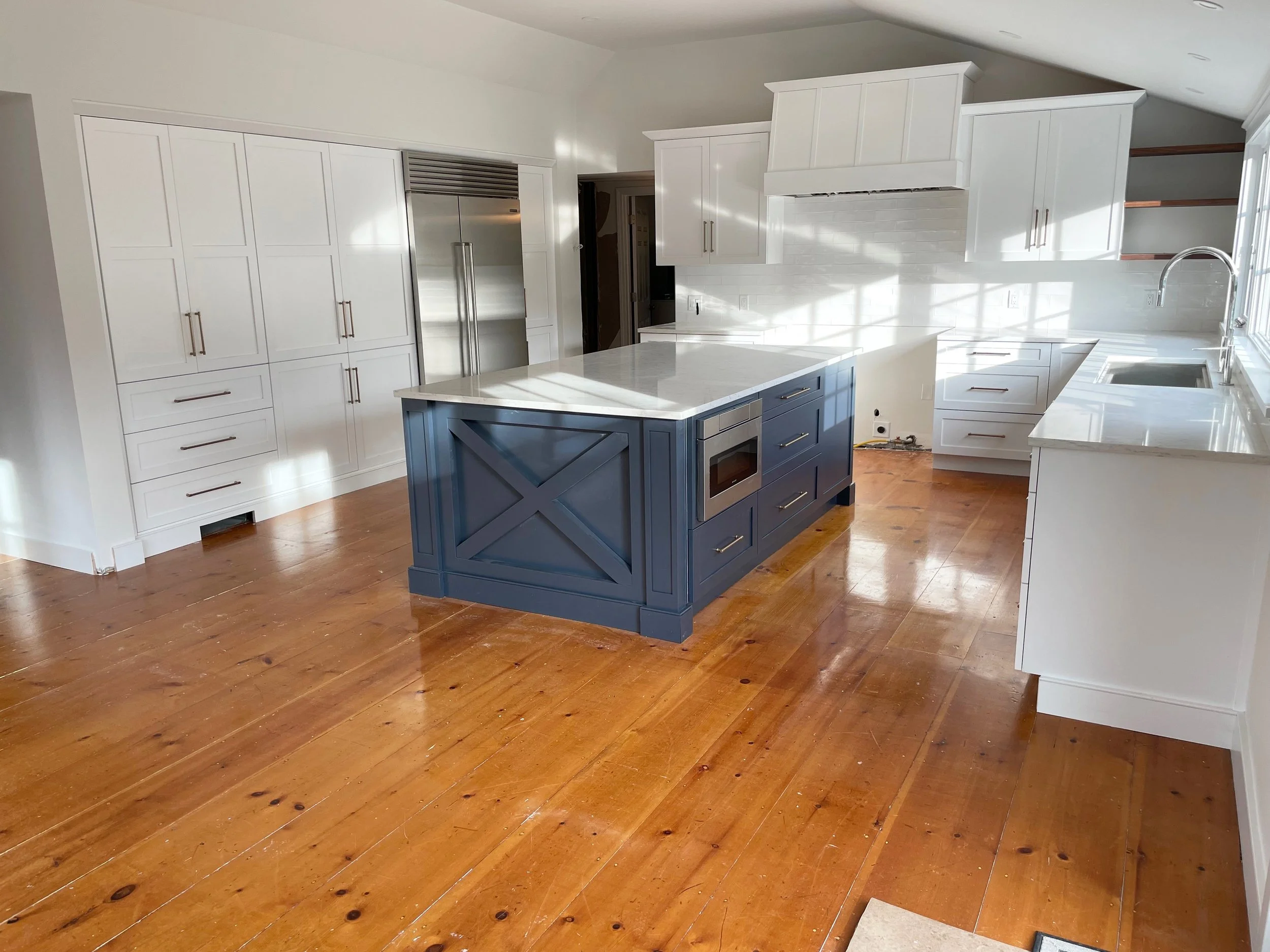 Hardwood floor installation in a Duxbury kitchen featuring a blue island, white cabinets, stainless steel appliances, and a marble countertop under natural light.