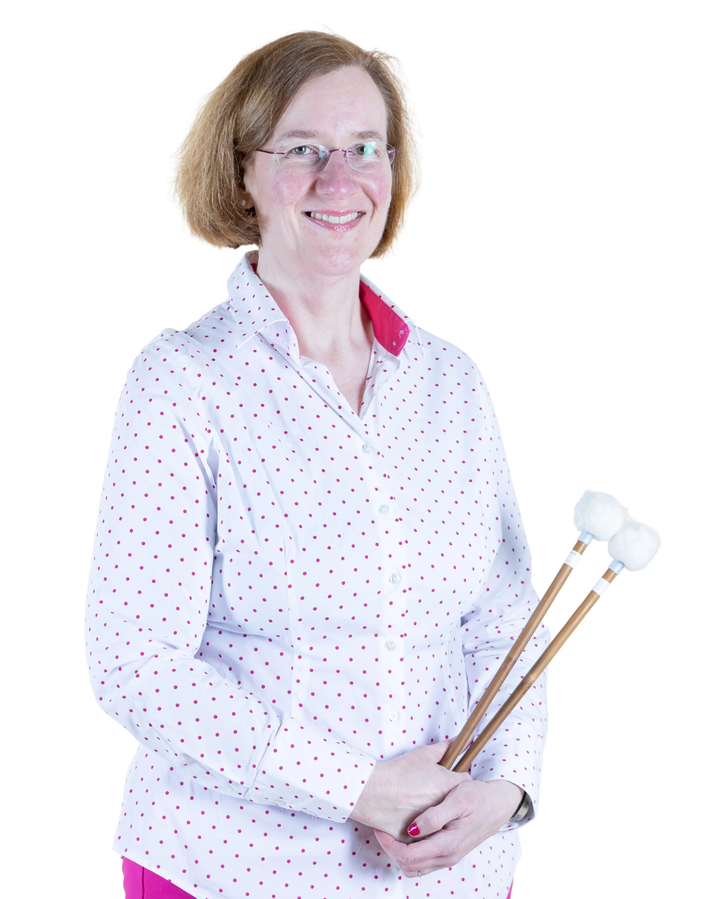 A woman with shoulder-length light brown hair and glasses, smiling, wearing a white button-up shirt with pink polka dots, holding two drumsticks wrapped in white cloth.
