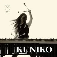 Female DJ playing turntables, holding a baton, with her hair blowing in the wind, and the name 'KUNIKO' on the equipment.