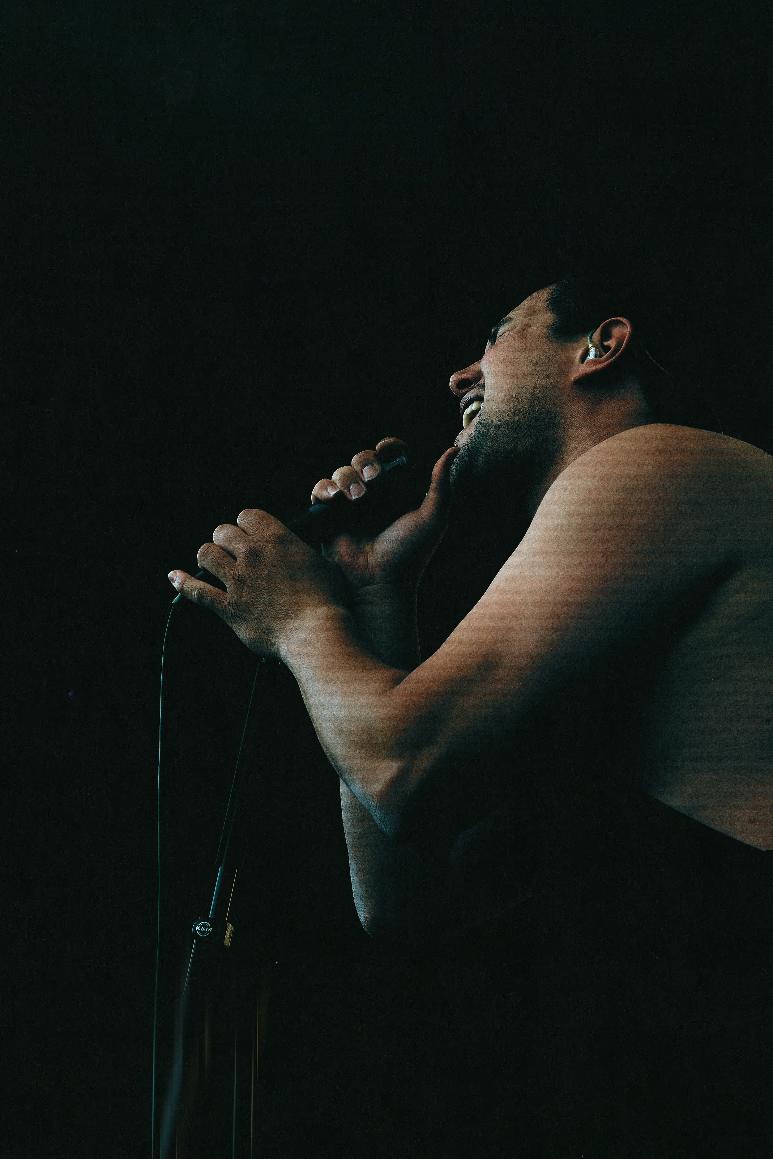 A man is holding a microphone close to his mouth, singing or speaking with a joyful expression on his face, against a black background.