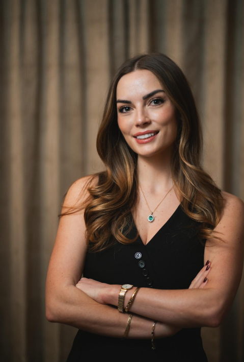 A woman with long brown hair and light skin is smiling, wearing a black sleeveless top, a necklace with a green pendant, and gold jewelry, standing in front of a brown curtain.