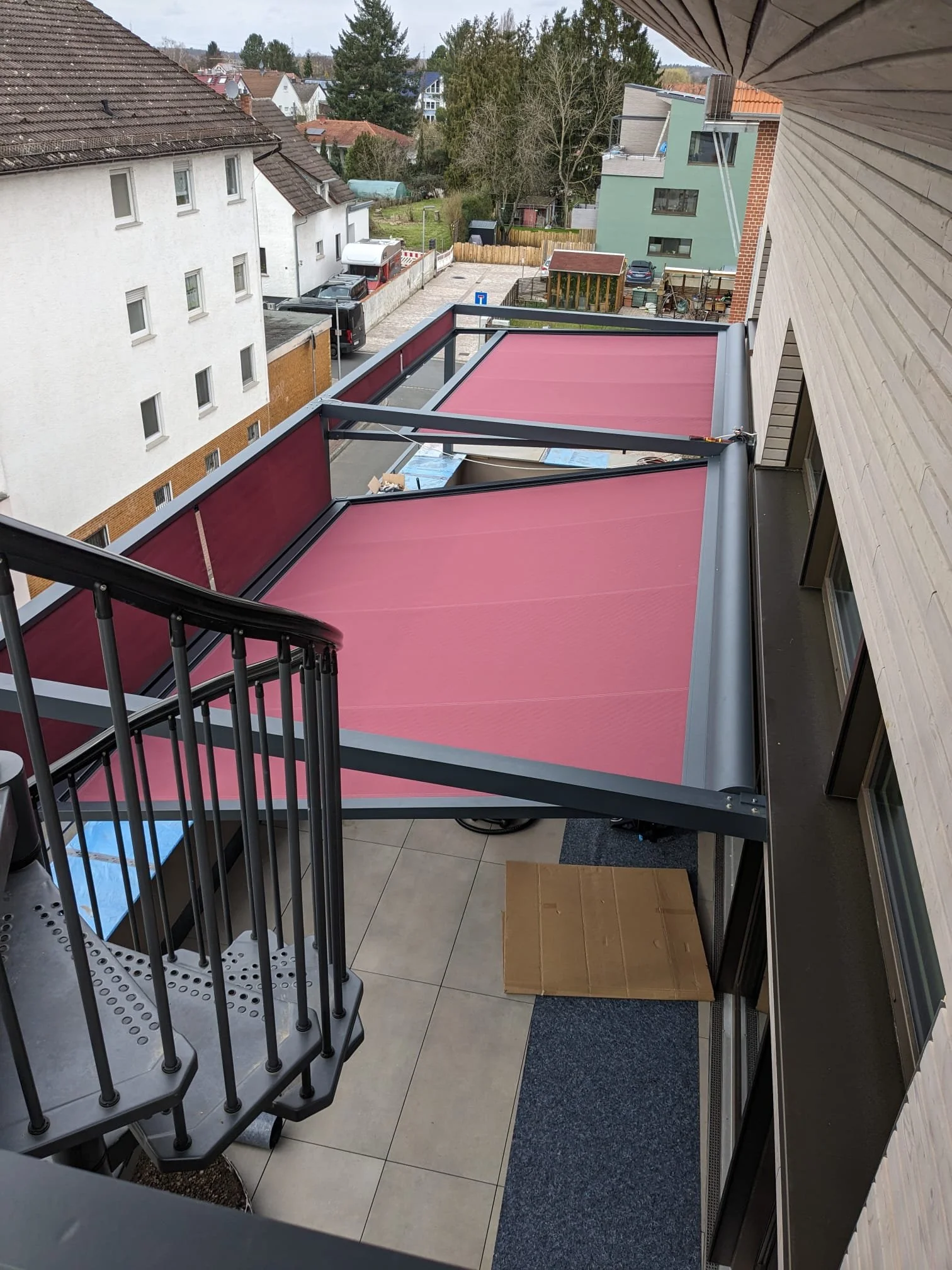 Blick von einem Balkon auf ein Mehrfamilienhaus mit einer Dachterrasse, Sonnenschutzmarkisen in rot, ein Treppenpodest mit Geländer, sowie umliegende Häuser, Straßen und einen Garten im Hintergrund.