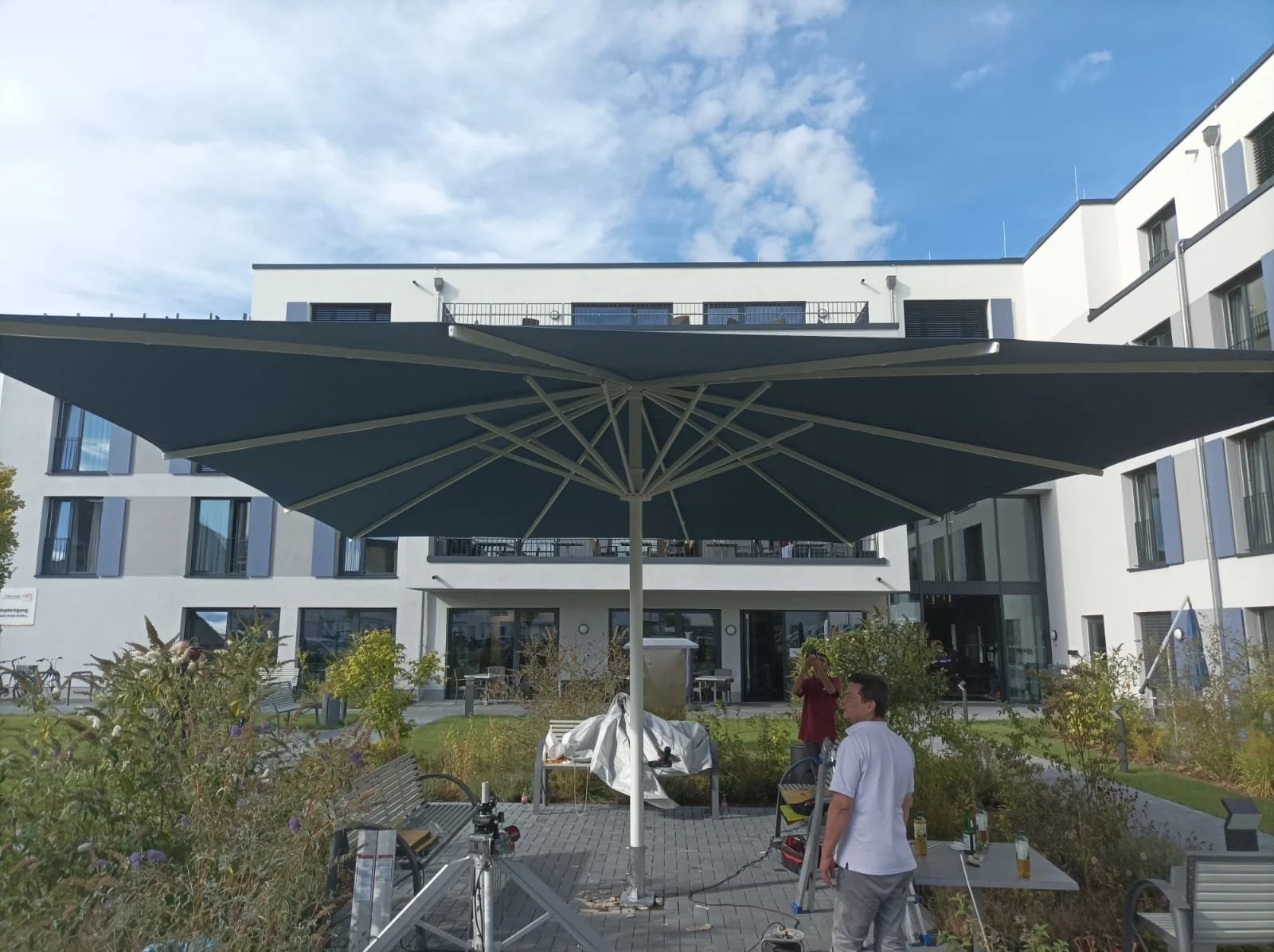 Ein moderner, weißer Mehrfamilienhaus-Komplex mit Balkonen. Im Innenhof steht ein großer Sonnenschirm, umgeben von Bänken, Pflanzen und Menschen, die an einem Film- oder Foto-Dreh beteiligt sind. Es gibt Kameras, Lichter und andere Ausrüstung.