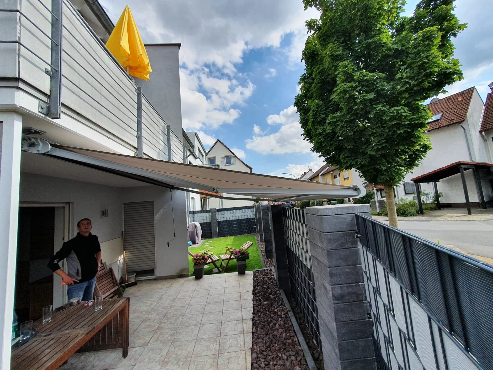 Blick auf eine Terrasse mit einem Mann, Gartenmöbeln, Pflanzen, einem Baum und einem Wohnhaus in einem Vorort bei schönem Wetter.