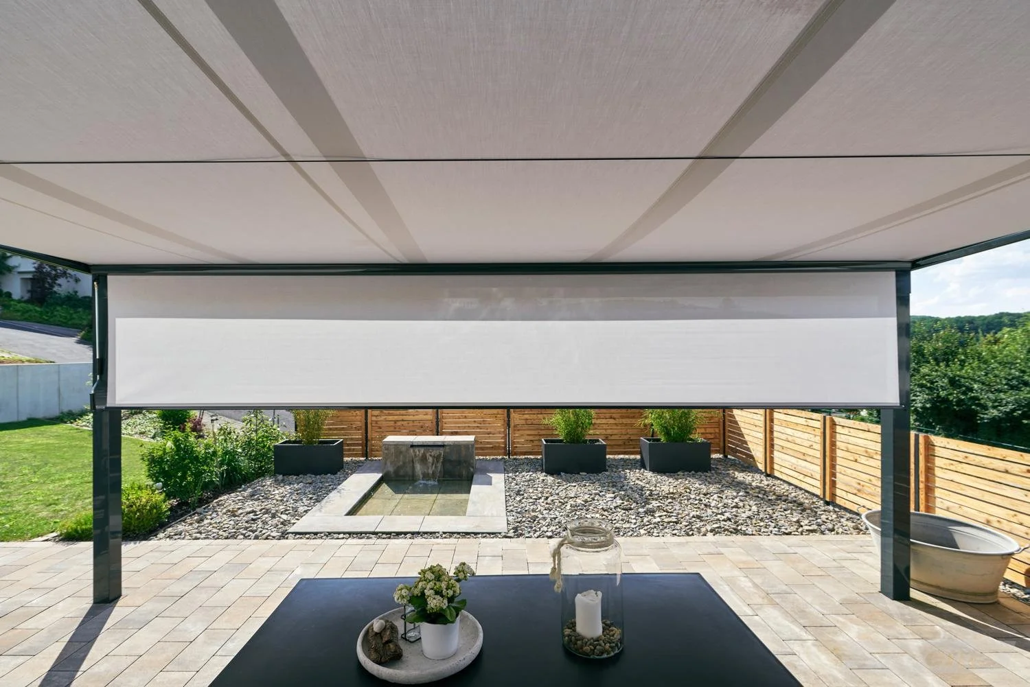 Überdachter Außenbereich mit Blick auf Garten, Wasserbecken und Pflanzen, sonniger Tag.