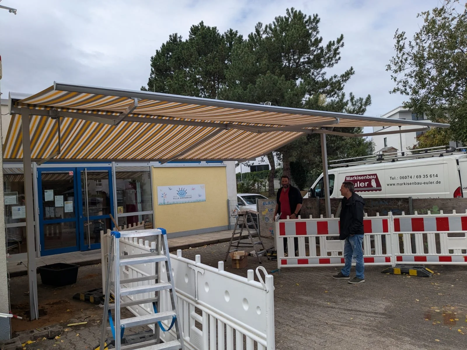 Auf Baustelle befindet sich ein gelber Eingang mit blauen Fenstern und einem blauen Rahmen. Es gibt eine Bauabsperrung mit weißen und roten Streifen, zwei Männer unter einer gelb-weiß gestreiften Markise, einen Leitern und einen weißen Kübel.