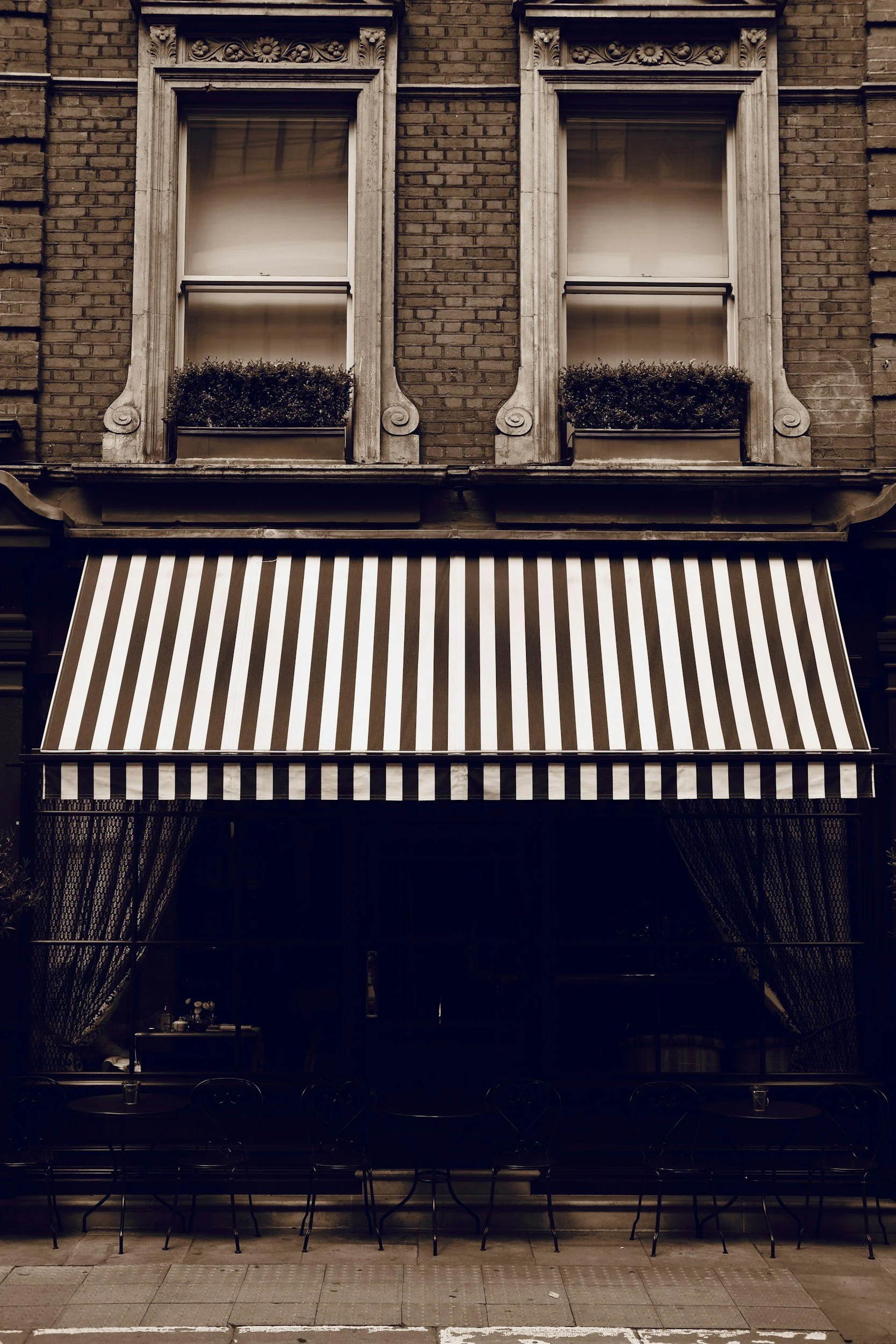 Ein zweistöckiges Gebäude mit zwei Fenster im oberen Stock, die jeweils Pflanzen in Fensterkästen haben. Das Erdgeschoss hat einen Laden mit einem schwarzen und weißen gestreiften Markisen. Vor dem Laden befinden sich mehrere schwarze Metallstühle und Tische.