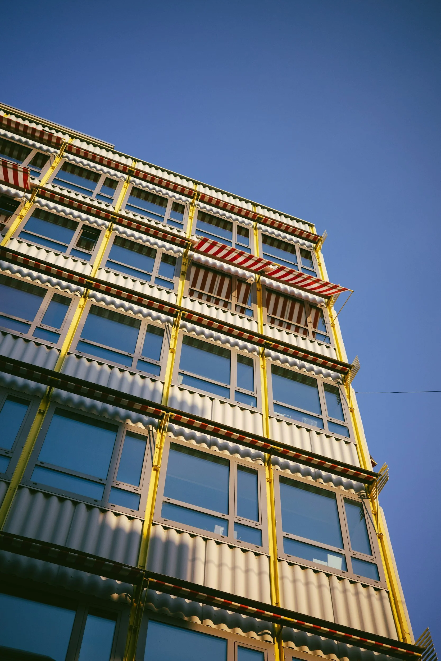 Blick auf ein modern gestaltetes Mehrfamilienhaus mit mehreren Geschossen, gelben Rahmen und großen Fenstern, unter einem blauen Himmel.