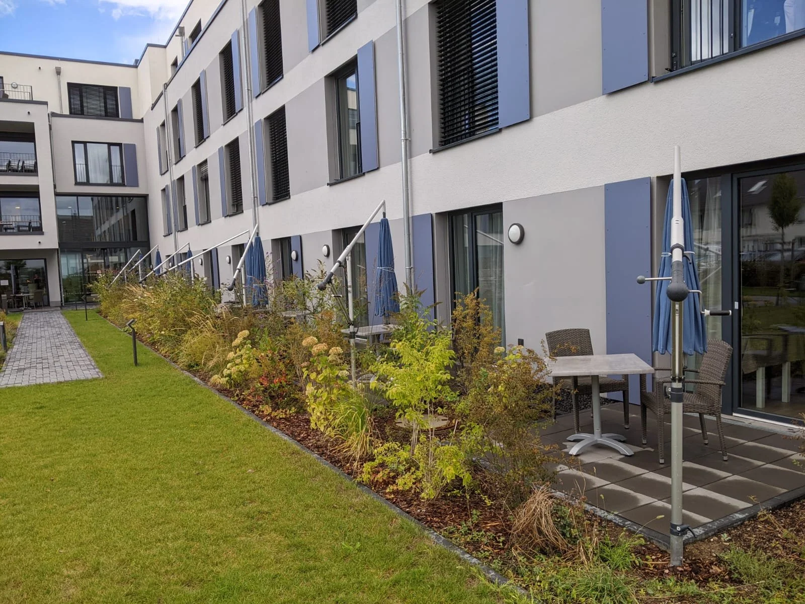 Ein moderner Gebäudekomplex mit mehreren Balkonen und großen Fenstern. Vor dem Gebäude befindet sich eine grüne Rasenfläche, angrenzend an einen gepflasterten Weg. Eine Terrasse mit Gartentisch, Stühlen und geschlossenen Sonnenschirmen ist sichtbar.