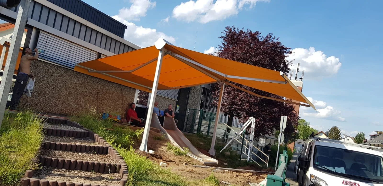 Kinder spielen auf einer Rutsche im Außenbereich mit Sonnenschutzunterstand. Ein Kind trinkt Wasser, während andere auf einer Bank sitzen und sprechen. Es ist ein sonniger Tag mit blauen Himmel und einigen Wolken.