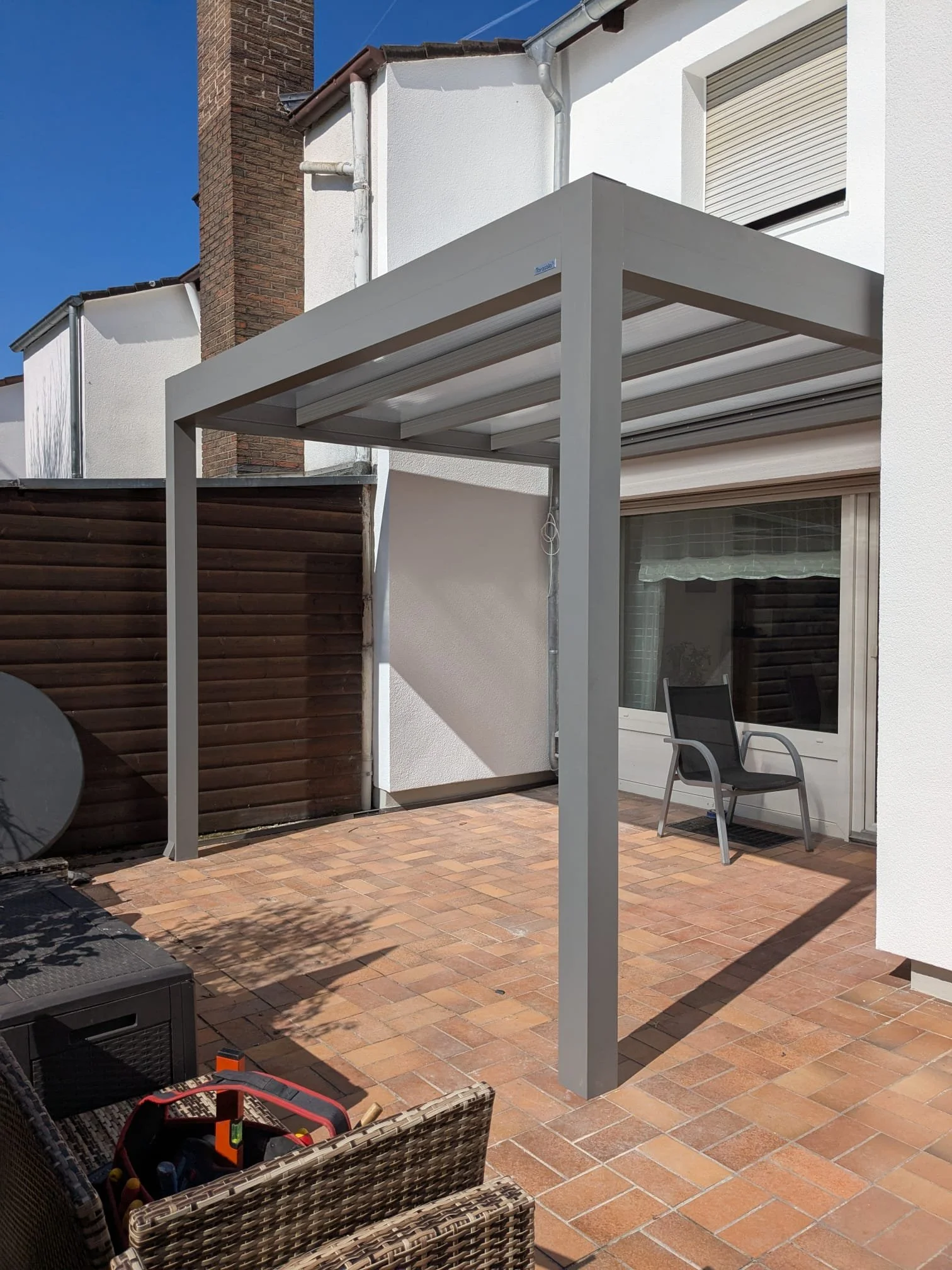 Ein moderner Patio mit überdachtem Bereich, sitzender Stuhl vor einem weißen Haus mit großen Fenstern und einer Markise, umgeben von einem Zaun aus Holz und weißen Wänden.