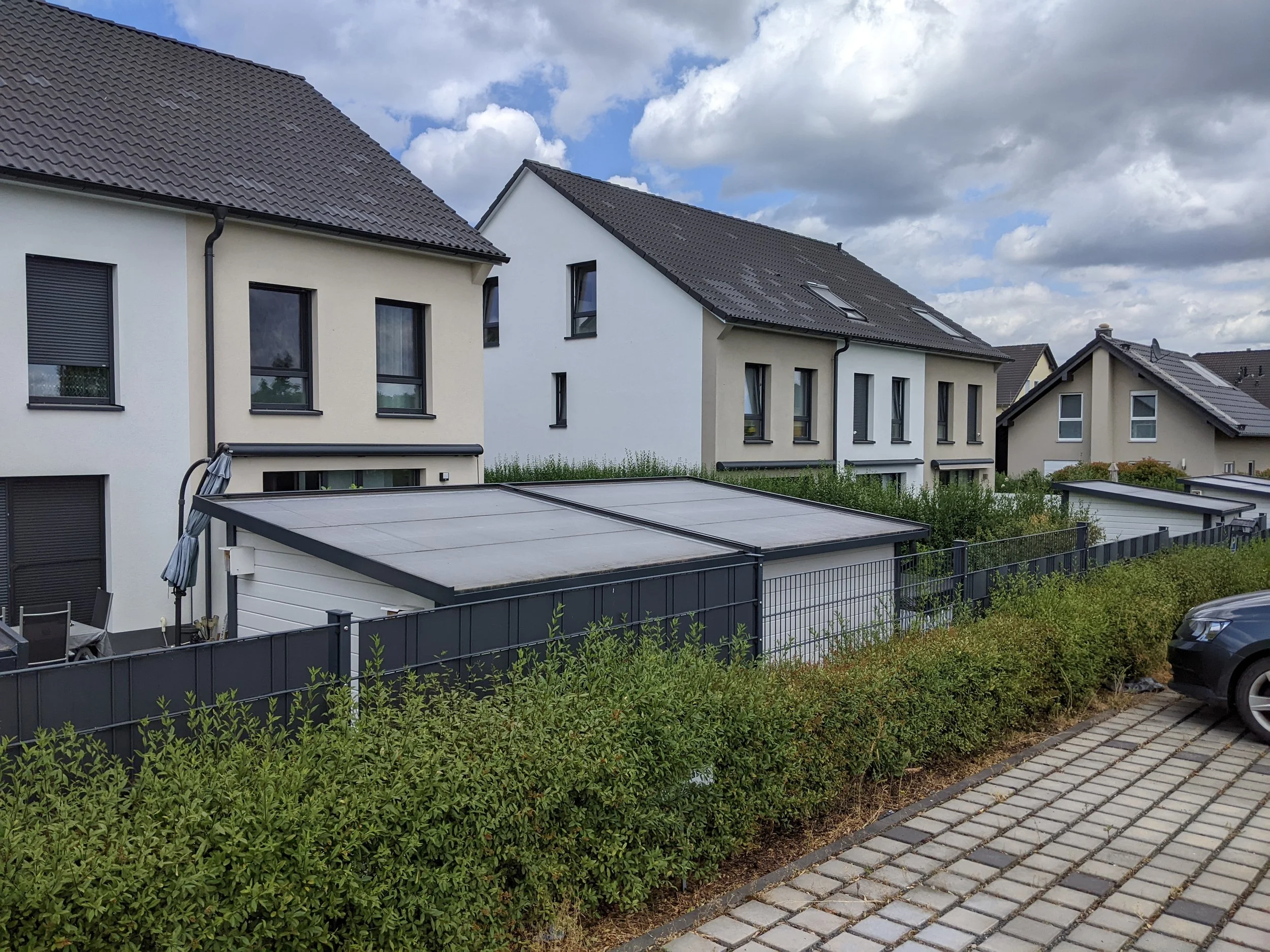 Mehrfamilienhaus mit braunen und weißen Wänden, schwarzem Dach und Dachfenstern, im Vordergrund eine Terrasse mit einem Sonnenschirm, einer umrandeten Hecke und einem Auto auf dem Parkplatz.
