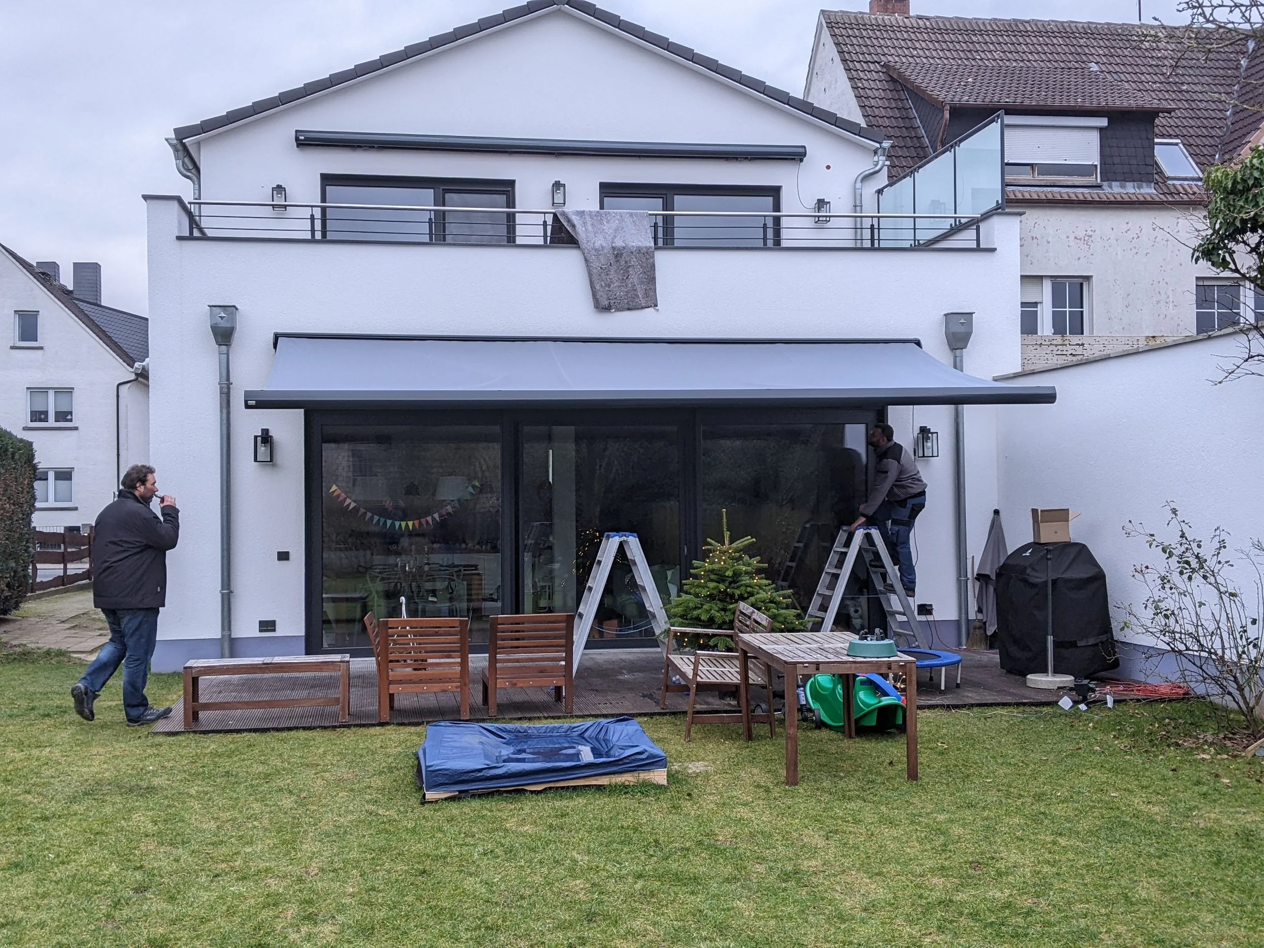 Drei Männer im Garten vor einem modernen weißen Haus; zwei Männer bauen zusammen vor der Terrasse, einer steht mit einem Getränk in der Hand, während der andere auf eine Leiter steigt, um eine Weihnachtsdekoration aufzustellen.