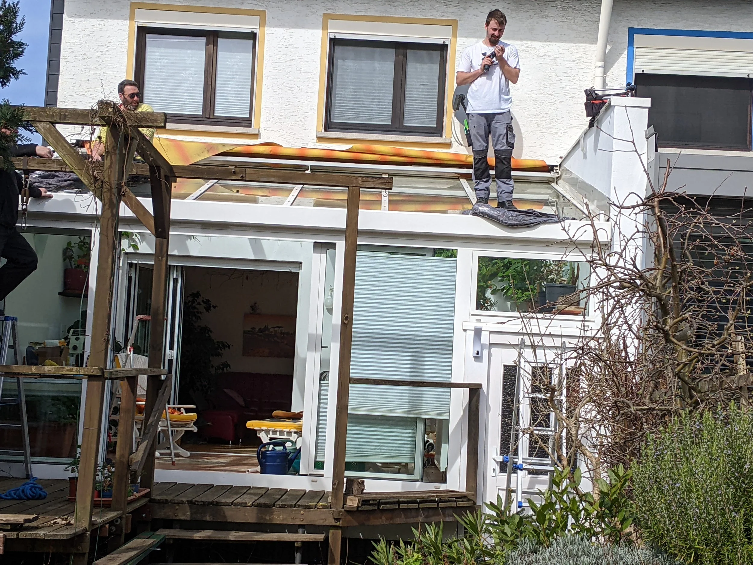 Zwei Männer arbeiten auf dem Dach eines Hauses, eines steht auf dem Dach und benutzt ein Werkzeug, das andere steht auf einer Leiter. Das Haus hat große Fenster und eine Terrasse darunter.
